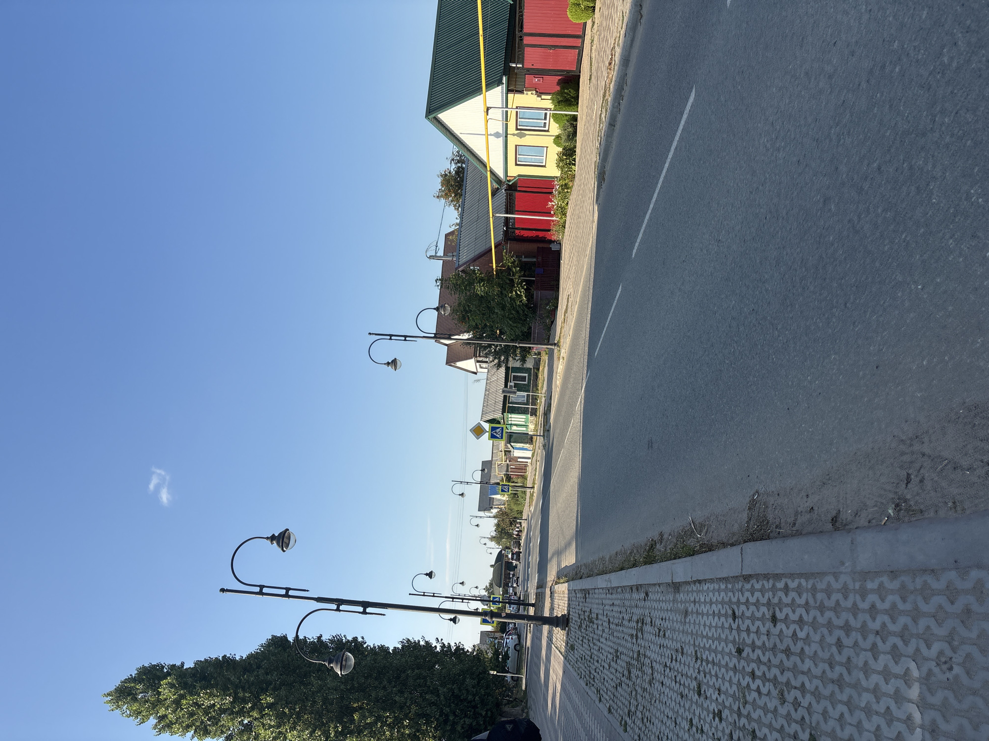 A street scene in Sol-Iletsk, Russia, under a clear blue sky. A paved road runs diagonally from the lower right to the upper left of the frame. To the left of the road, a concrete sidewalk is laid with patterned pavers, bordered by a grassy verge and then a row of streetlights with curved arms. Beyond the sidewalk, a tall, dense green tree stands on the left. On the right side of the road, several houses with colorful facades and pitched roofs are visible. The houses have red and yellow paint, with red fences. Traffic signs, including a yellow diamond-shaped sign and a pedestrian crossing sign, are placed along the roadside. A few parked cars and some distant figures on the street suggest a quiet daytime setting. The overall environment appears to be a residential street in a town or small city.