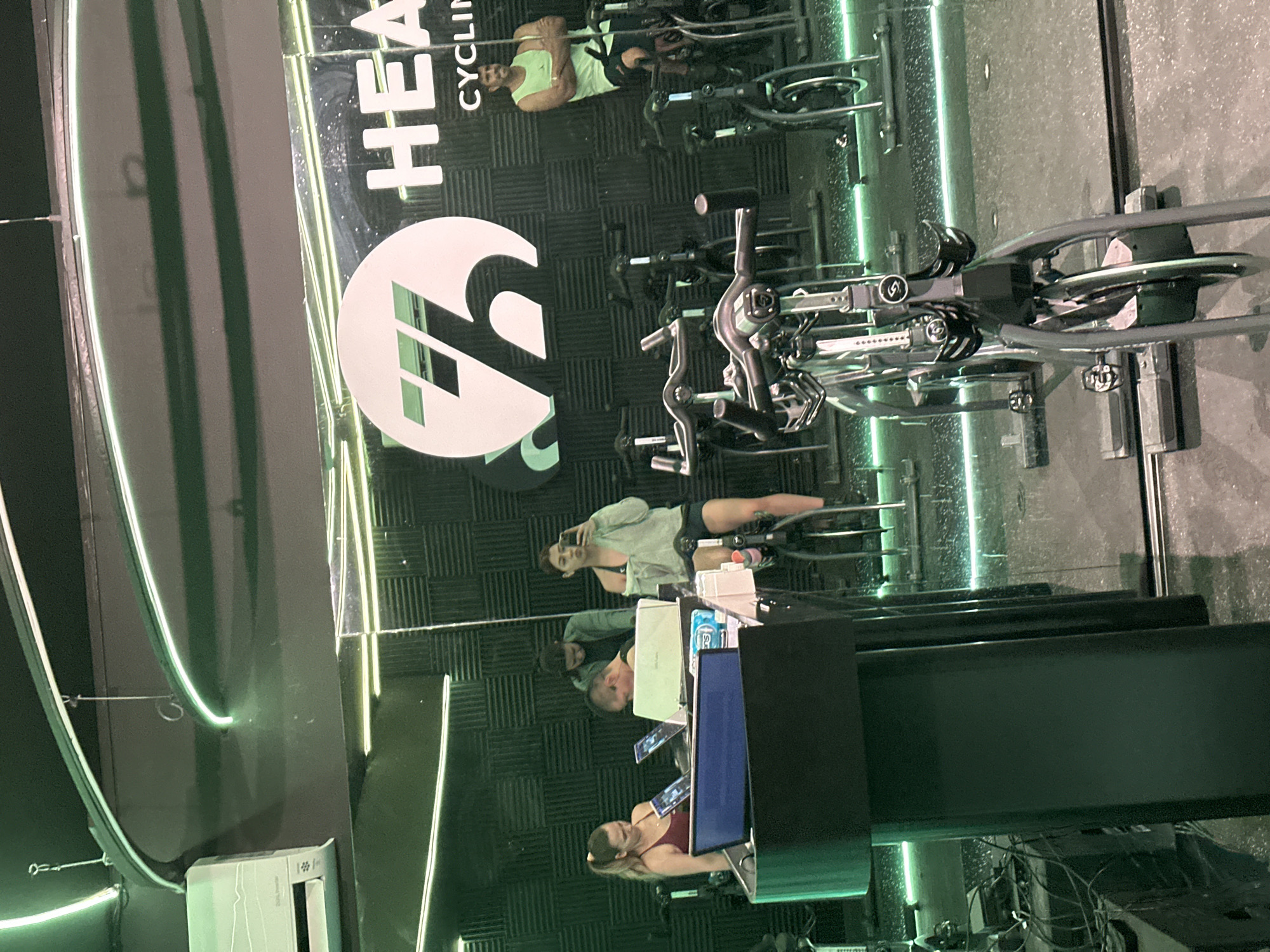 The image depicts an indoor cycling studio, likely in Puebla, Mexico, with a mirror reflecting the studio's interior. Several stationary bikes are arranged in rows. People are visible on the bikes, appearing to be engaged in a cycling class. In the foreground, a desk or counter with monitors and equipment is present. The studio has a dark interior with green accents and a large "HEAT Cycling" logo on the wall.