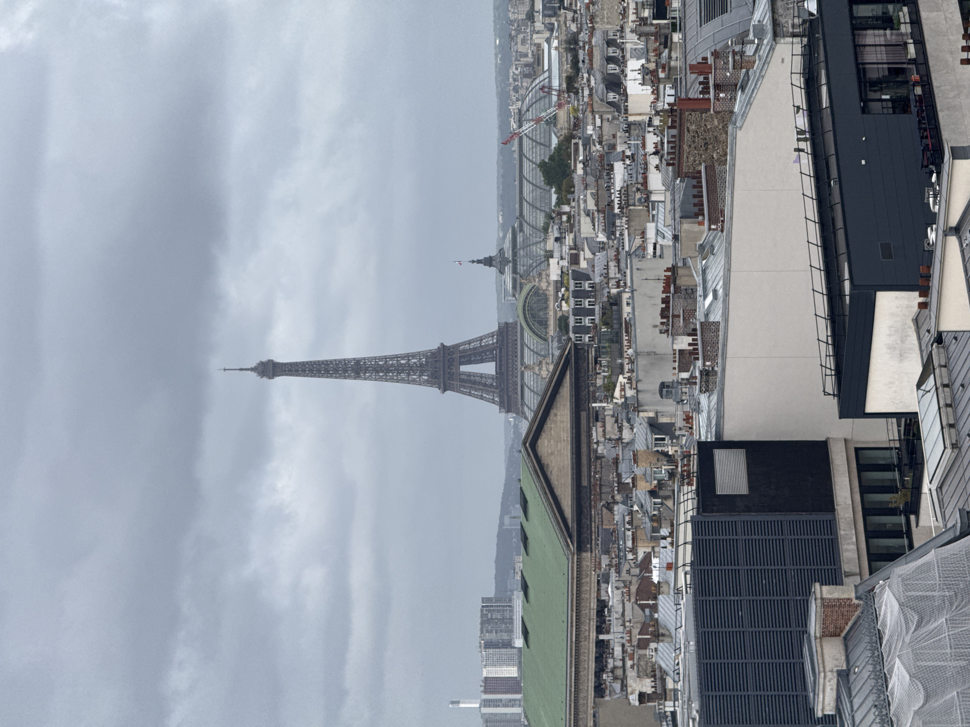 The image captures an elevated view of Paris, France, featuring the Eiffel Tower prominently in the background. The foreground reveals a dense urban landscape of rooftops and buildings, suggesting a high vantage point within the city. Overcast skies create a soft, diffused light, casting a somewhat muted tone across the scene.