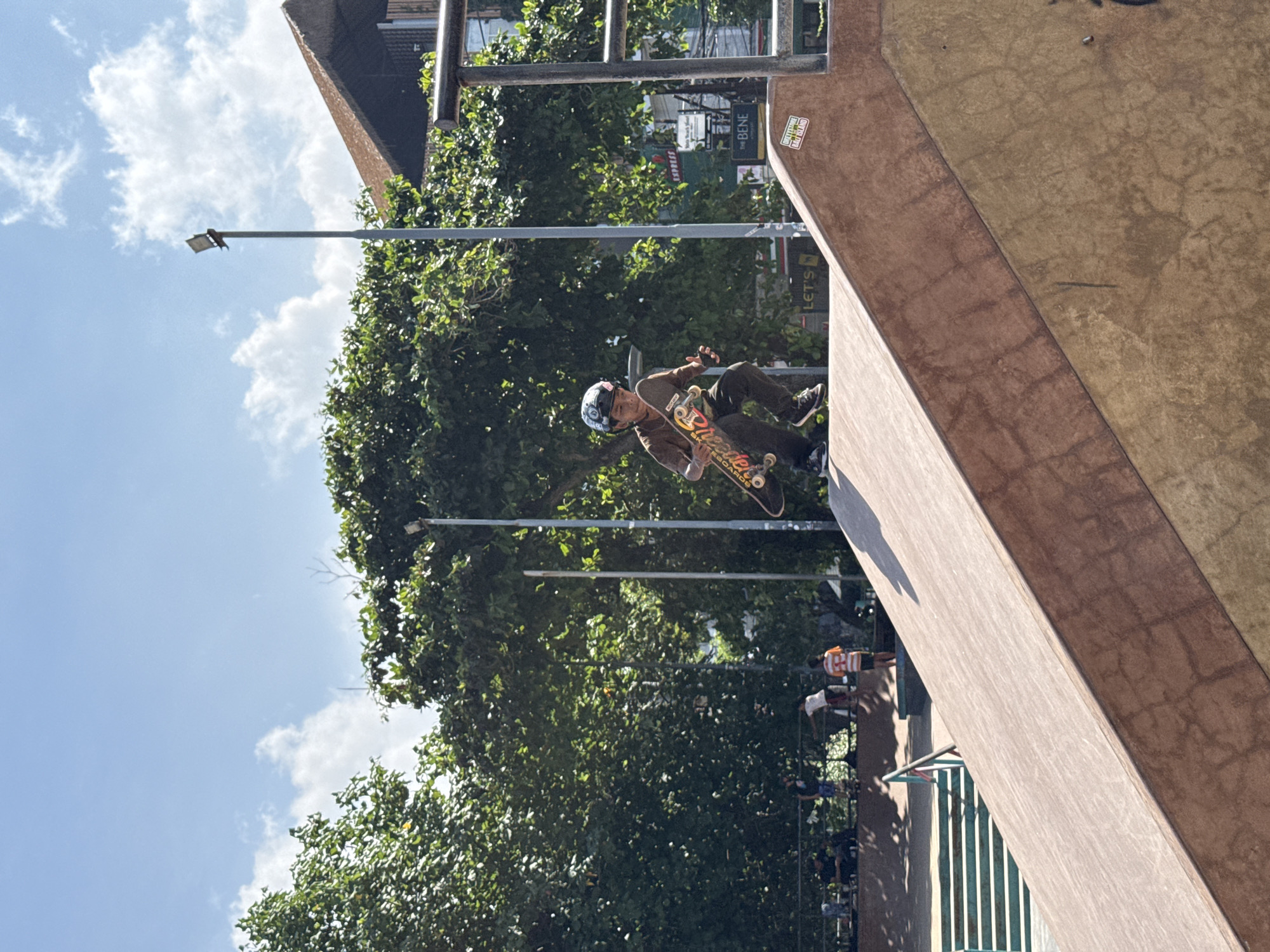 The image depicts a skateboarder in action at a skatepark located in Kuta, Indonesia. The skateboarder, wearing a helmet and casual clothing, is airborne, seemingly executing a trick on a ramp. The ramp is constructed of plywood and is situated outdoors. In the background, there is lush greenery with trees, streetlights, and partial views of buildings. There are a few people observing the skateboarder's performance.