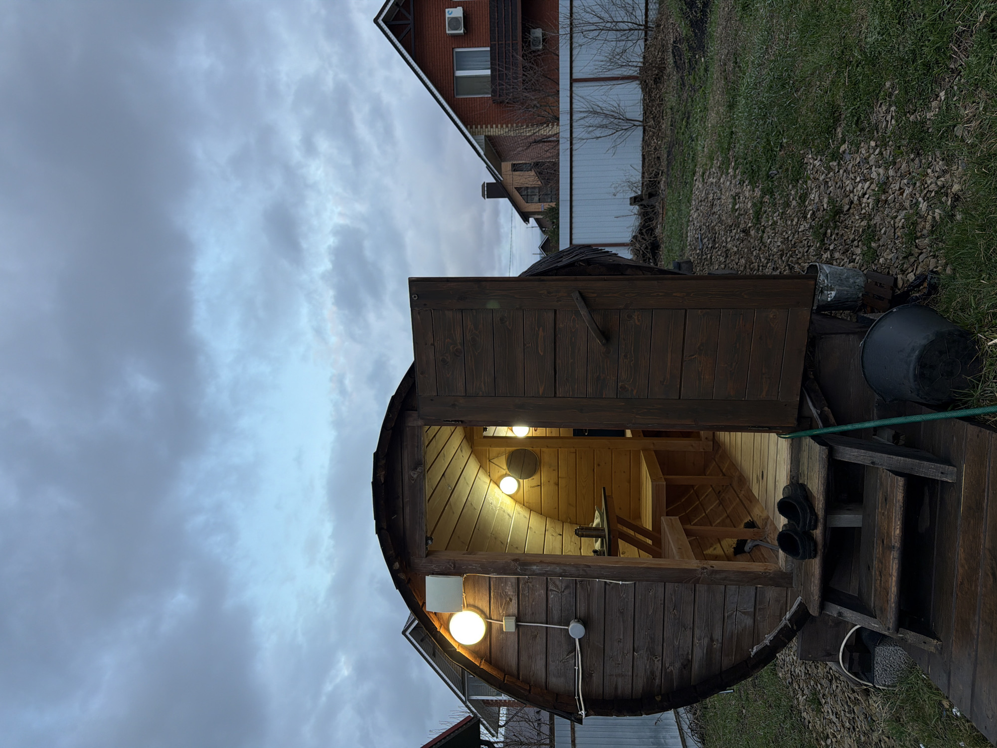 The image depicts a barrel-shaped sauna in what appears to be the backyard of a property located in Большая Шильна, Russia. The sauna is constructed of wood, with an open door revealing an illuminated interior. Inside, there are wooden benches and a table. Externally, the sauna has exterior lights, and a small wooden staircase leads to the entrance. A green hose is visible near the sauna. In the background, there is a house with a red brick exterior, along with a white fence and some vegetation. The sky is overcast.