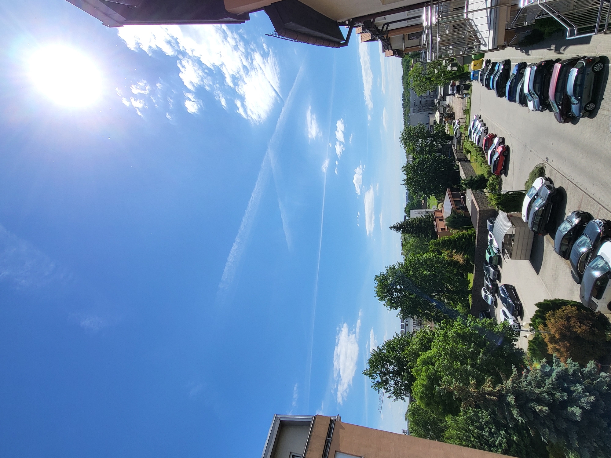 This image shows a view from an elevated position, likely a building. The scene is dominated by a bright blue sky with contrails and scattered clouds. Below, a row of parked cars lines the edge of a street, which is bordered by trees and buildings. The buildings are a mix of residential and possibly commercial structures. The overall impression is that of a typical urban or suburban street scene on a sunny day.