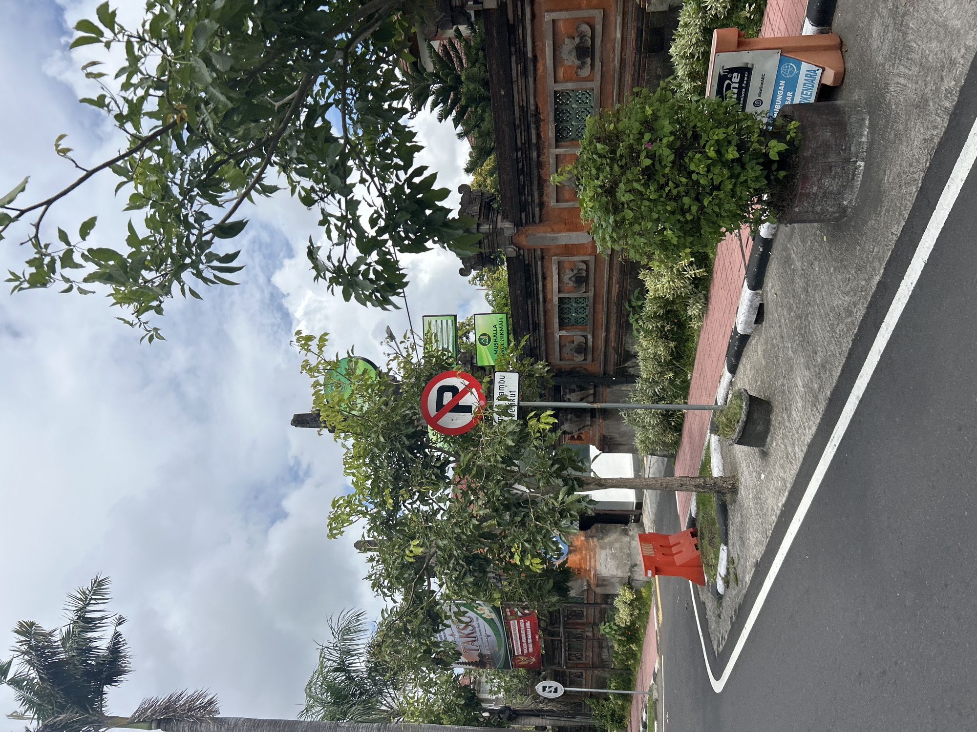 In Denpasar, Indonesia, an urban street scene unfolds under a cloudy sky. A road with a white dividing line curves to the left, bordered by a red brick sidewalk with black and white curbs. Orange traffic barriers and a small tree with a concrete planter sit on the side of the road. Beyond the sidewalk, a decorative Balinese stone wall, adorned with intricate carvings and green latticed windows, stretches across the frame. Lush green trees and palm trees frame the upper portions of the image, obscuring parts of the sky. Several signs are visible: a "no parking" sign with a red circle and a diagonal line through a blue "P," a sign with Indonesian text reading "TAMBU" and "SEKUT," a "TAKSO" banner, and other green rectangular signs with text in Indonesian. A traffic sign with a white "E" in a blue circle is also present. The overall impression is one of a street in a tropical city, likely in a tourist-heavy area given the decorative wall and signage.