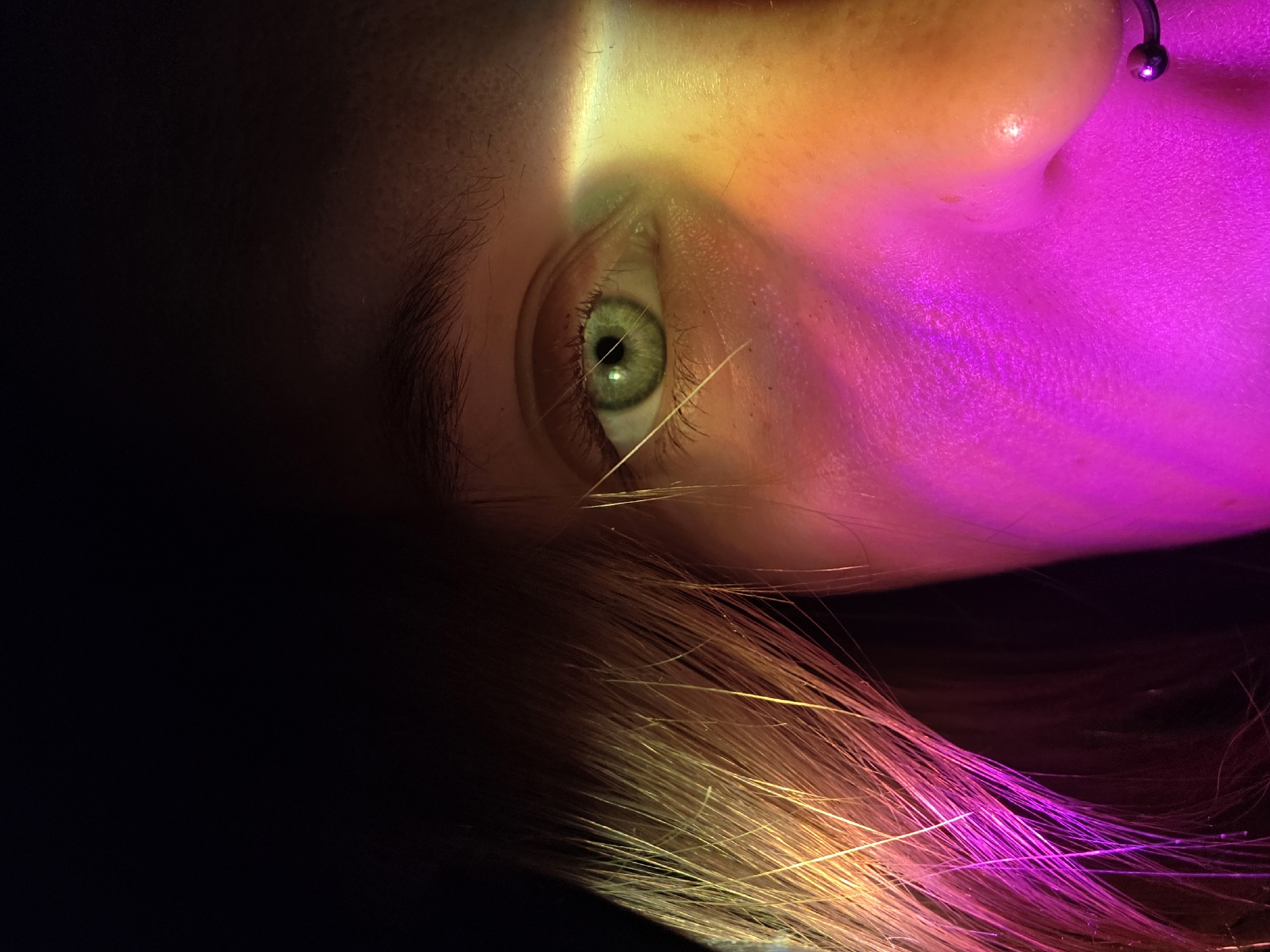 The close-up image shows a person's eye, nose, and hair, illuminated by a beam of colored light that casts pink, yellow, and orange hues across the face. The eye is green, with a dark pupil. A septum piercing is visible on the person's nose. The hair, which appears to be long and light-colored, is also bathed in the colorful light. The background is predominantly black, creating a stark contrast with the illuminated portion of the face. No specific location in Minsk, Belarus, can be determined from the image.