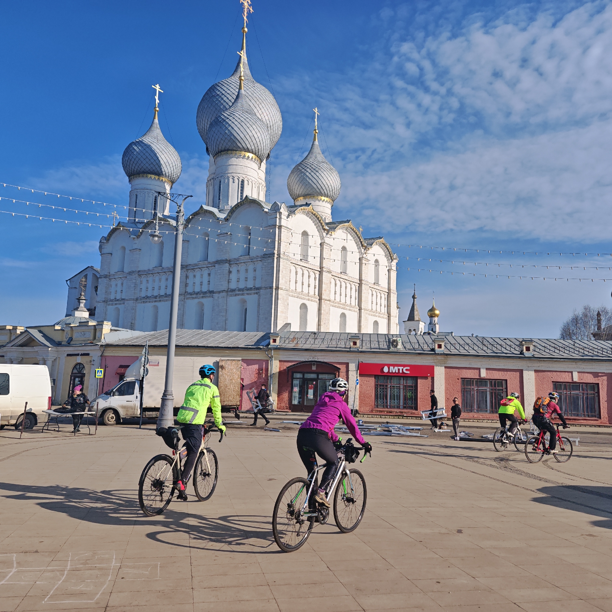 The image depicts a daytime outdoor scene in Rostov Veliky, Russia, featuring the Assumption Cathedral (Uspensky Cathedral) within the Rostov Kremlin under a bright blue sky with scattered clouds.
In the background, the prominent white stone cathedral rises with five grey, fluted, onion-shaped domes, each topped with a golden cross. The central dome is significantly larger than the others. Decorative arched windows and facade elements are visible. To the right and below the cathedral, a lower, reddish-brick building with multiple rectangular windows is present, one section bearing a red "MTC" (MTS) sign. Another section of this building displays a pink facade. To the far left, a yellow-colored building with a small dome and cross is visible. Strings of white lights are horizontally strung across the upper portion of the mid-ground. A black lamppost stands near the center-left.
In the mid-ground and foreground, three individuals are riding bicycles from left to right across a paved square. The lead cyclist on the left wears a neon yellow-green jacket and a blue helmet, riding a light-colored gravel bike. The middle cyclist wears a magenta jacket and a white helmet, riding a silver/grey gravel bike with green accents. The third cyclist, on the right, wears dark clothing with an orange-accented backpack and a helmet, riding a red gravel bike. Their long shadows are cast on the light-colored pavement, where chalk hopscotch markings are visible in the foreground.
Other individuals are present in the mid-ground. Near the MTC building, one person in dark attire carries a large, light-colored rectangular panel horizontally, while another person stands nearby. To the far left, near a parked white panel van and a silver/grey truck, a person wearing a pink hat is seated at a white table. A yellow pedestrian crossing sign is visible near the truck.