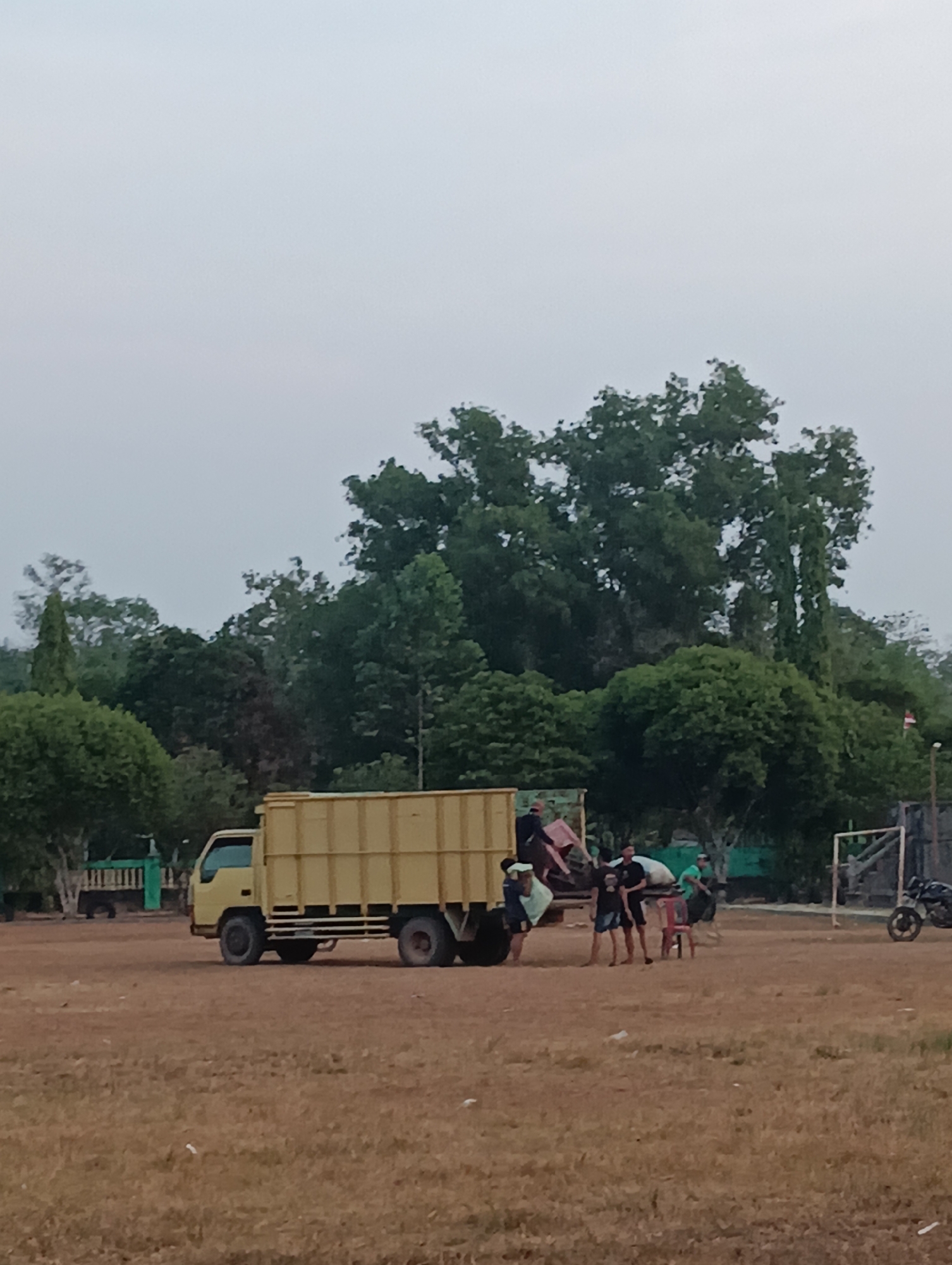 In Pematang Jaya, Indonesia, a group of individuals are gathered around a yellow truck on a dusty, outdoor field. The truck, a box truck with a light yellow cargo area, is parked at an angle. Several people are near the back of the truck, with some appearing to be unloading or arranging items from its bed. One person is visible inside the truck's cargo area, and others are standing on the ground near it. A motorcycle is parked to the right of the scene, near a metal structure that resembles a goalpost. The background is filled with lush green trees under a pale, overcast sky. The ground is covered in dry grass and dirt.