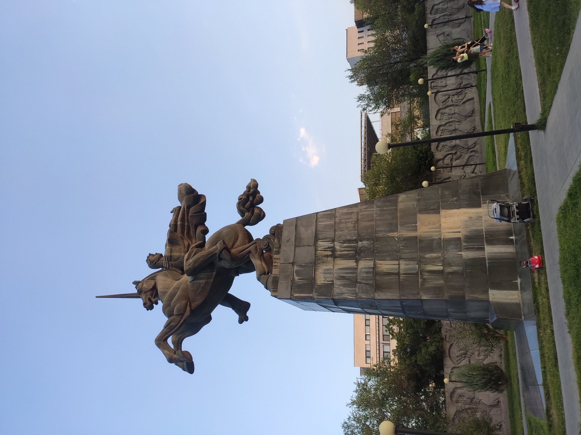 The image captures the equestrian statue of David of Sassoun in Yerevan, Armenia. The statue depicts David, a legendary Armenian hero, on horseback with a sword raised. It stands on a massive stone pedestal in a park-like setting. In the background, modern buildings and trees are visible under a clear blue sky. Several people are present in the park, including a family with a stroller and children on scooters. A red toy car is on the grass near the base of the statue. The scene appears to be set in the late afternoon or early evening, with soft lighting.