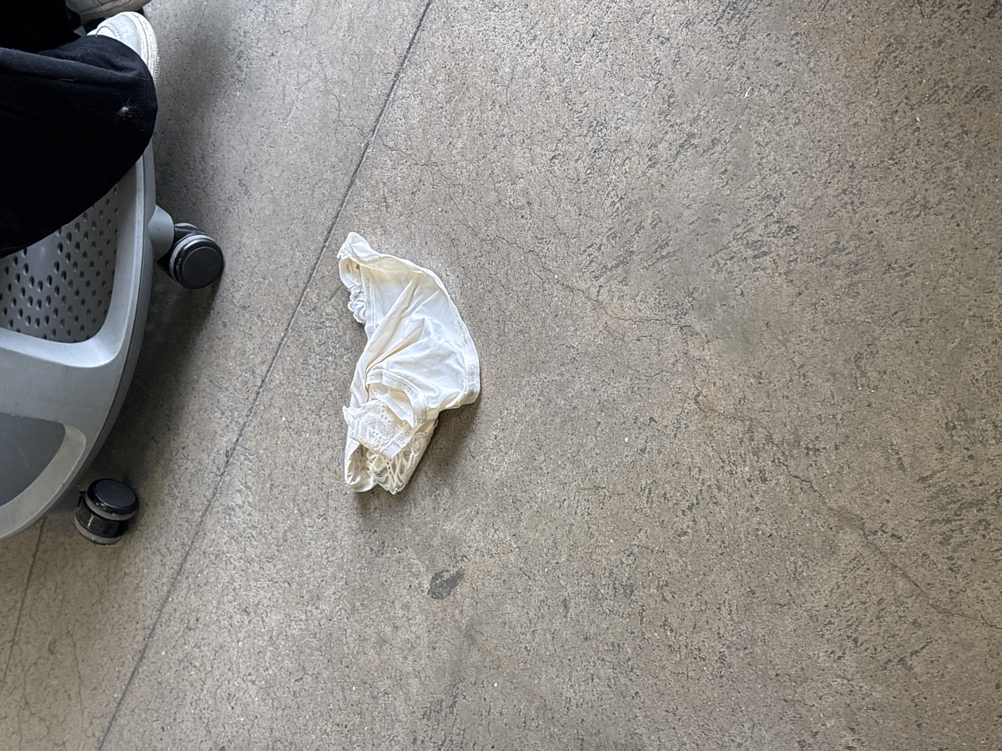 A white, lace-trimmed undergarment lies on a gray concrete floor, near a wheeled office chair. A person wearing black pants and white sneakers is seated in the chair. The location appears to be an interior space with a plain concrete floor. There are no other people or objects clearly visible. The scene's setting cannot be definitively identified as Mexico City.