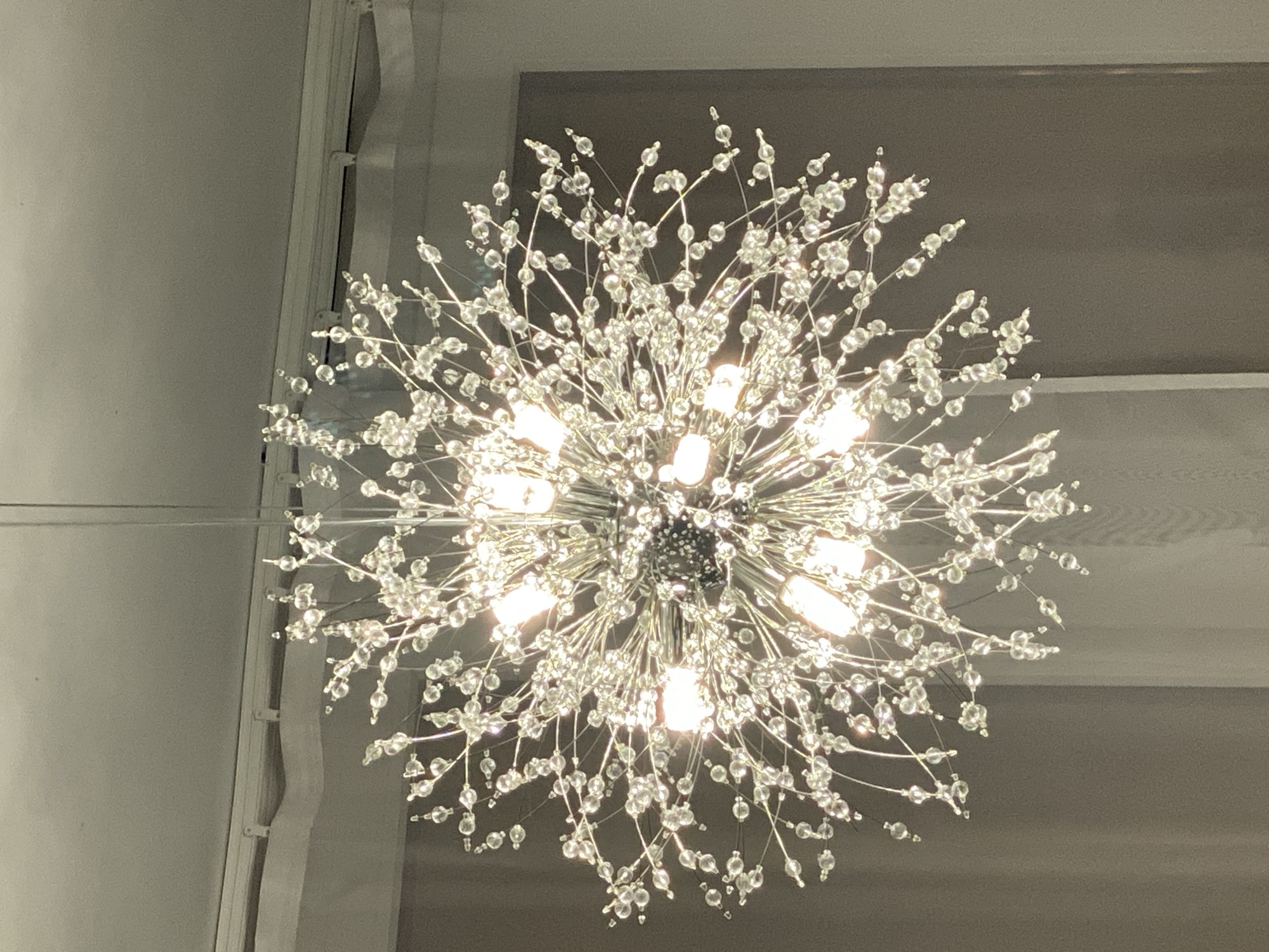 A close-up shot captures a sparkling, dandelion-shaped chandelier hanging from a ceiling. The light fixture is adorned with numerous small, clear beads and appears to be made of a silver-colored metal. Several bulbs are illuminated, casting a bright glow that reflects off the beads, creating a dazzling effect. The background consists of a textured ceiling and hints of sheer curtains or drapery, suggesting an indoor setting. The location is unknown.