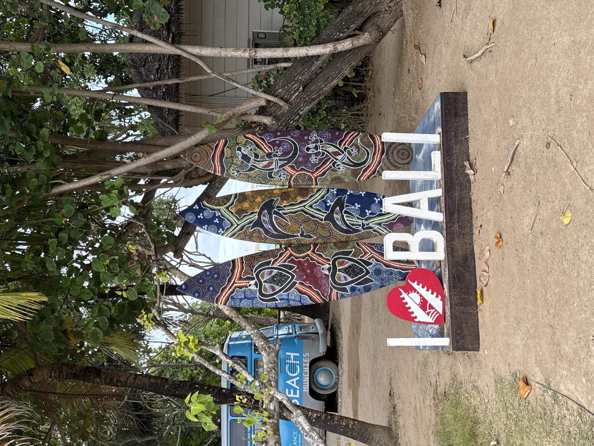 An outdoor scene in Kuta Selatan, Indonesia, features a set of three vibrantly decorated surfboards standing upright behind a large, white "I LOVE BALI" sign. The surfboards display intricate Aboriginal-inspired artwork featuring turtles and lizards in black, red, yellow, and blue patterns. The "I LOVE BALI" sign is composed of large, white block letters, with the "I" and the heart symbol for "LOVE" being the most prominent in the foreground, followed by "BALI." The heart is red and adorned with a white sun and stylized waves. The sign is placed on a dark wooden base. To the left of the surfboards, a blue and grey van with "BEACH MUNCHIES" and "BEACH RESORT" written on its side is parked on a sandy surface. The background includes lush green trees, some with palm fronds, and a glimpse of a white building with a thatched roof, characteristic of tropical resort architecture. The ground is sandy and scattered with fallen leaves and twigs. The overall setting suggests a beachside tourist location.
