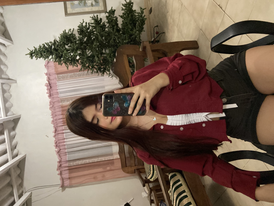 A young woman poses in a mirror selfie. She has long brown hair and is wearing a red jacket over a white top, paired with dark shorts. She holds a phone with a sparkly case up to her face, partially obscuring it. The background includes a Christmas tree, light-colored walls, a window with pink curtains, and a chair. The overall impression is casual and stylish.