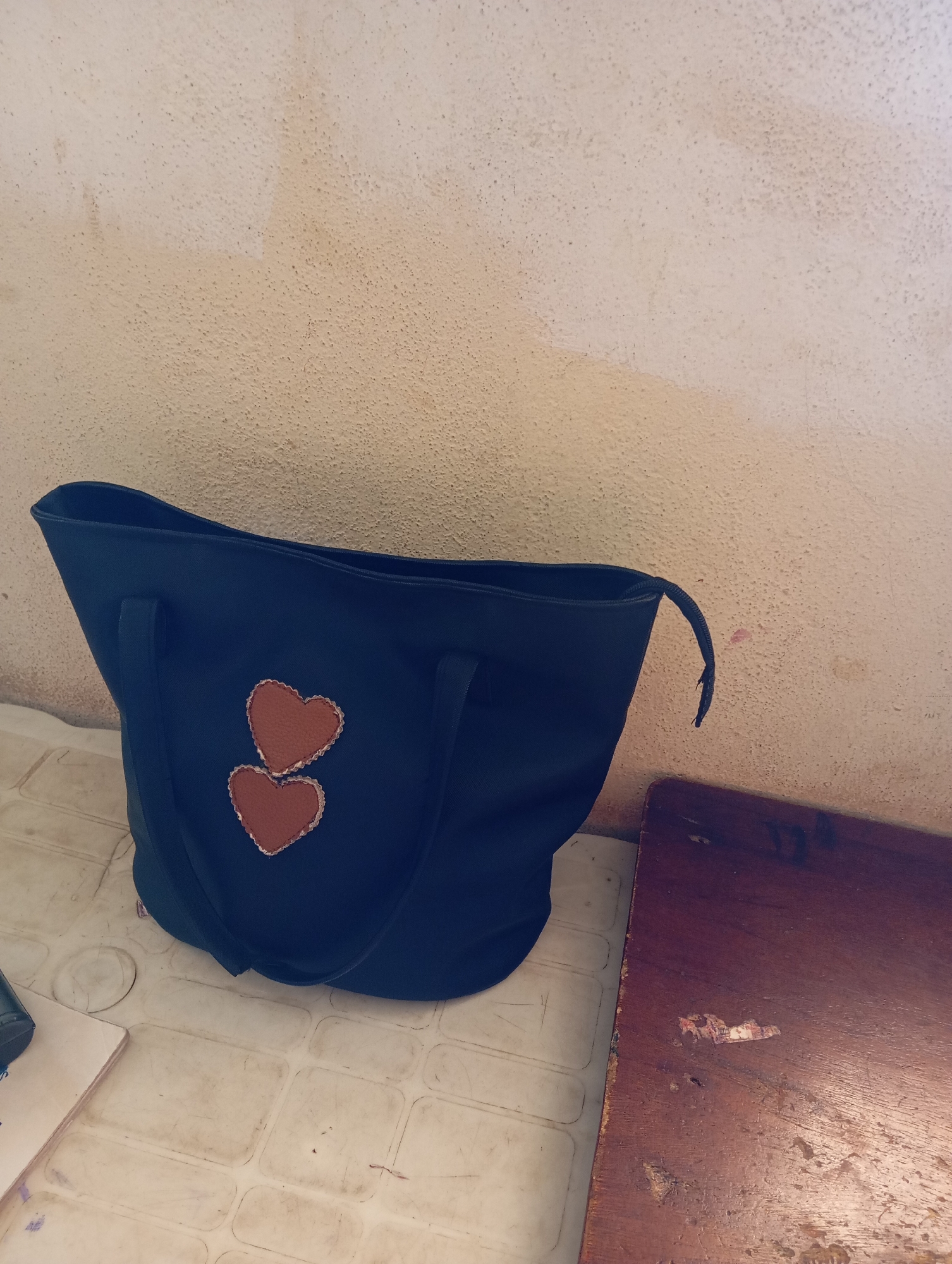 A dark blue tote bag with two stitched orange heart appliques sits on a textured, off-white surface, possibly a table or floor, in Gura Top, Nigeria. The bag's handles are visible, and a loose strap extends from its side. Behind the bag, a light beige wall with a rough, textured finish is slightly discolored in patches. To the right of the bag, a portion of a dark brown wooden surface, likely a table or desk, is visible, showing signs of wear and some peeling remnants of what may be labels or stickers. The overall scene suggests an indoor setting.