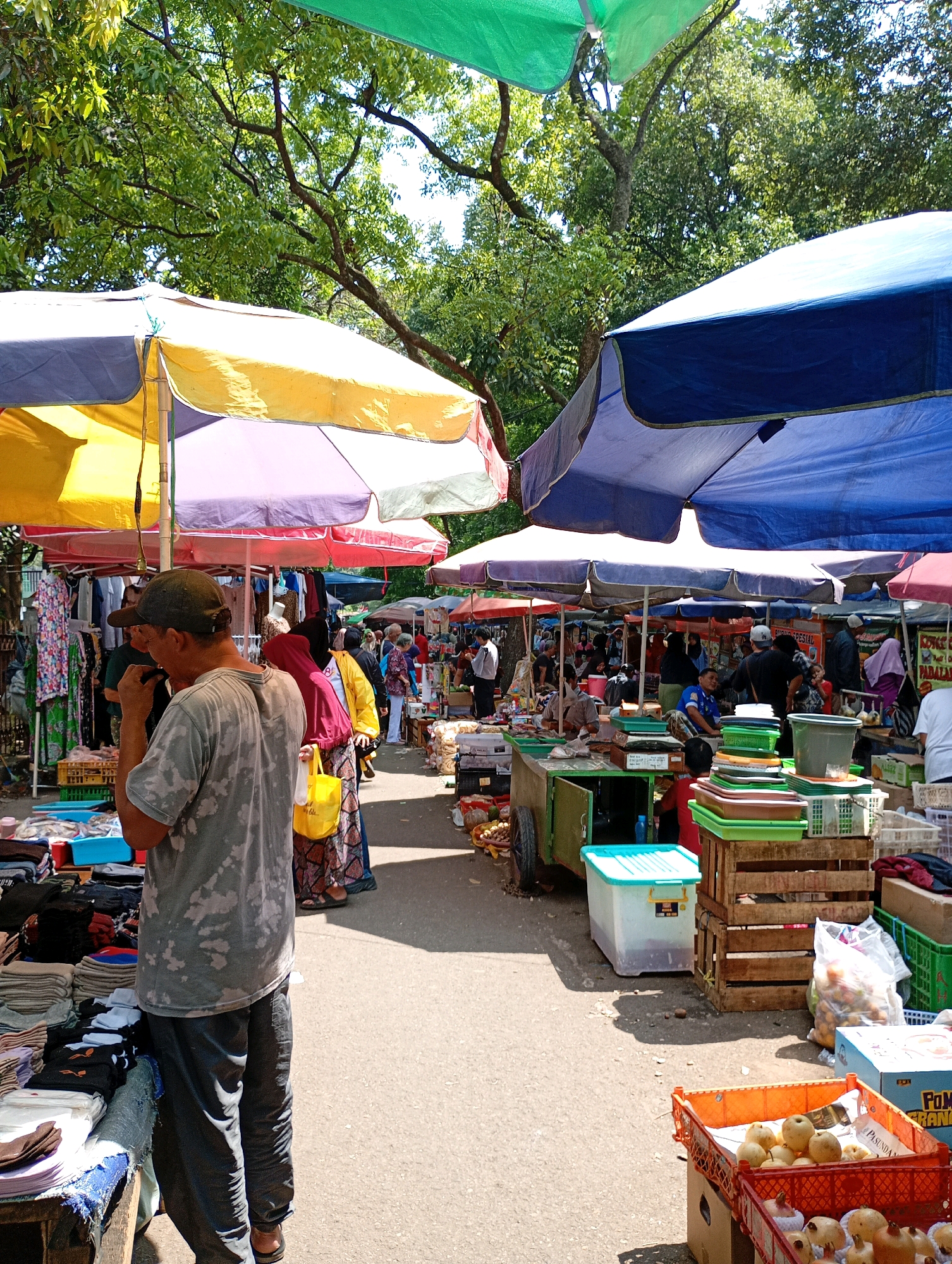 This image depicts an outdoor market scene, likely in Bandung City, Indonesia, given the context provided. The market is covered by numerous colorful tarps and umbrellas, including shades of green, yellow, purple, and blue, providing shelter from the sun. Stalls are set up along a paved pathway, displaying a variety of goods.
In the foreground, a man wearing a grey, tie-dye t-shirt and dark pants is seen from the back, standing near a stall. The stall features stacks of folded fabric or clothing in various colors and patterns. Adjacent to this, another stall displays more folded items, some in plastic bags.
Further into the market, other individuals are browsing or working at their stalls. A woman in a maroon headscarf and patterned skirt carries a yellow bag. Several stalls are laden with produce, including boxes and crates of fruits like pomegranates and pears. Various containers, baskets, and plastic tubs are also visible, used for storage and display. The overall impression is a bustling marketplace with diverse merchandise and shoppers. The environment appears to be daytime, with sunlight casting shadows on the ground. Trees with dense foliage are visible in the background, suggesting an open-air setting.