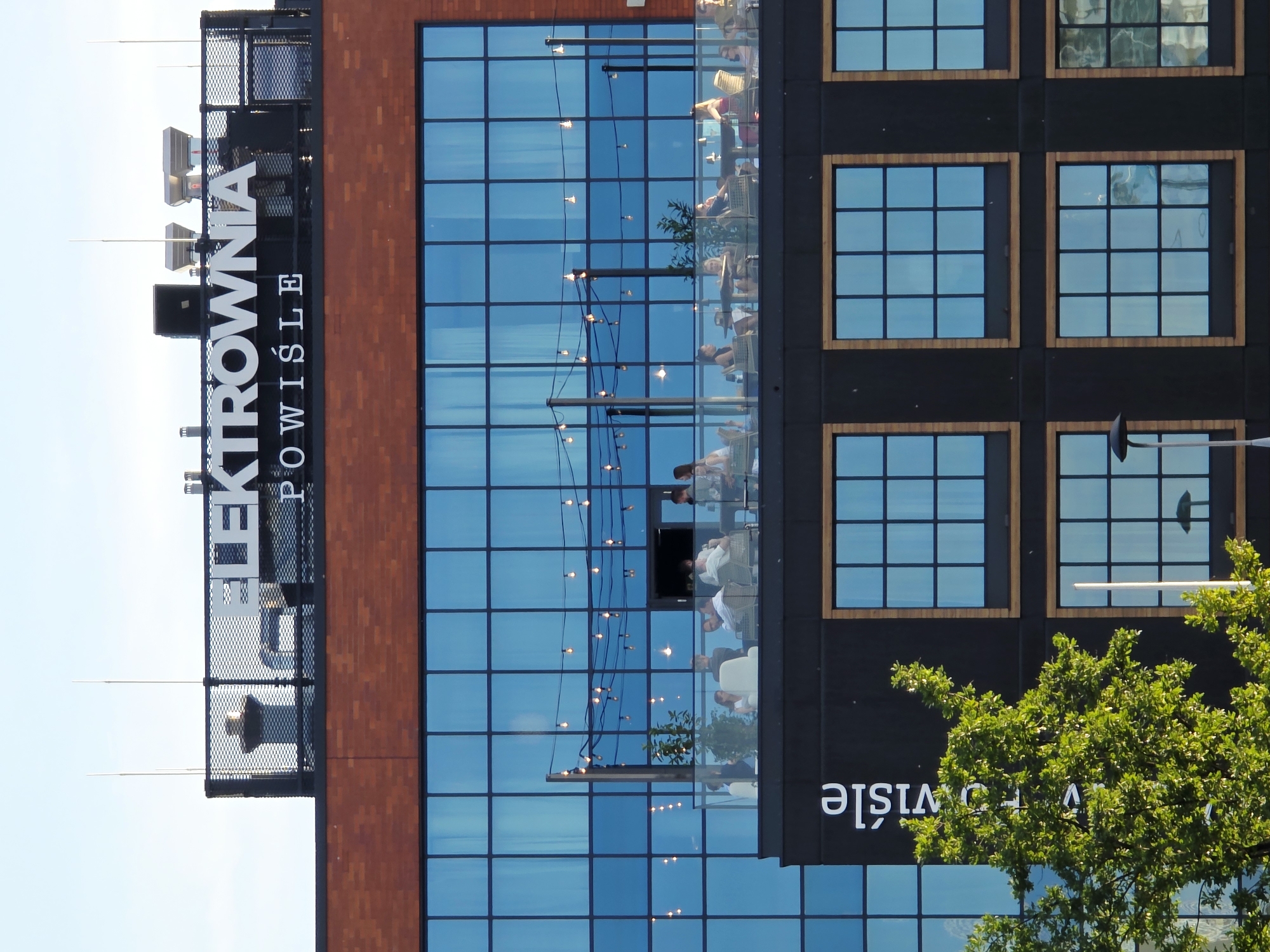 The image shows the exterior of a modern building in Warsaw, Poland, with the name "ELEKTROWNIA POWIŚLE" displayed prominently on its rooftop. The building features a brick facade on its upper section and a black facade with large, blue-tinted windows on its lower sections. A rooftop terrace is visible, populated with people dining and socializing at tables. String lights are suspended above the terrace, adding a decorative element. A lush green tree partially obscures the lower portion of the building, which has a repeating pattern of large, square windows. The sky above is a clear blue, indicating daytime.