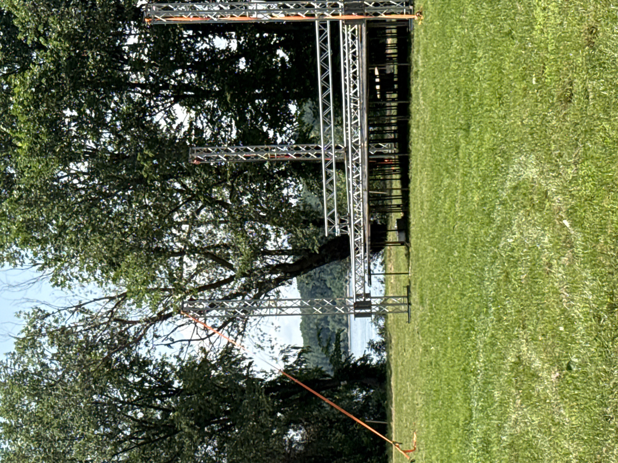 This image captures an outdoor scene, likely a field or park, set up for an event in Mechka, Bulgaria. The primary subjects are several metal truss structures, presumably part of a temporary stage, archway, or support system. These structures, made of silver-colored metal latticework, extend horizontally across the middle ground of the image. One section of the trussing appears to be a freestanding arch or support, while another segment, featuring a bright orange strap, is positioned closer to the foreground. The orange strap extends downwards, suggesting it's anchoring or marking a path.
The setting is characterized by a lush green grassy area in the foreground and to the right, indicating a well-maintained field. On the left and in the central background, numerous large trees with dense green foliage provide a natural backdrop. Beyond the trees and the truss structures, a wide body of water, possibly a river or lake, is visible, with a distant treelined shore or landmass beyond it under a clear sky.
The scene depicts preparations for an outdoor activity or event, given the temporary truss structures. There are no people visible, suggesting the event has not yet started or is being set up. The bright, clear lighting indicates it is daytime, likely a sunny day. No text is visible in the image. The overall atmosphere is calm and anticipatory, characteristic of an event venue before public access.