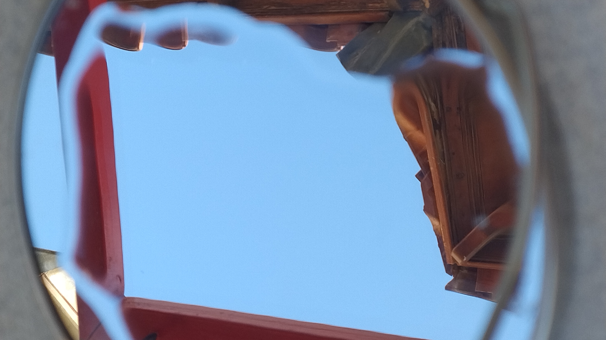 The image shows a reflection in what appears to be a circular or oval lens, possibly from eyeglasses or a camera lens. The reflection is distorted and fragmented, depicting a clear blue sky and sections of an outdoor structure. A portion of a red-painted beam is visible at the top left of the frame, running horizontally. Below this, there is a curved, lighter colored element, likely part of the framing of the lens. The lower half of the reflection shows details of a weathered, terracotta-tiled roof, with wooden beams and trim visible. A hint of greenery, possibly a plant or foliage, is also present on the right side, against the wooden structure of the roof. The scene's setting in Perea, Greece, suggests an outdoor environment, possibly near the sea or a building with a distinctive architectural style.