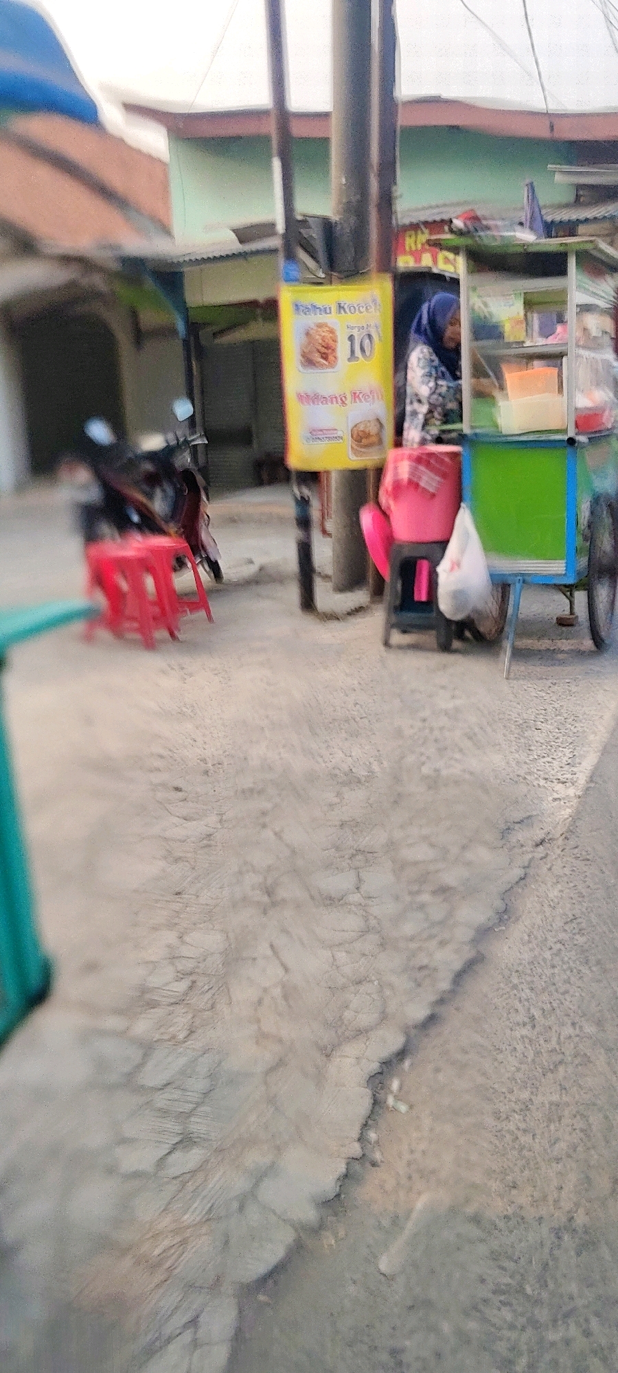 The image shows a street scene in Mekarmukti, Indonesia. In the foreground, the cracked and weathered pavement is visible. A red plastic stool is positioned on the pavement, with a motorcycle parked behind it. Further back, a food cart is set up, with a woman wearing a blue hijab standing behind it. A yellow sign on the cart displays text and a price of 10. Several plastic containers with food items are visible inside the cart. A pink plastic stool and a white plastic bag are placed next to the food cart. The background shows buildings and a utility pole.