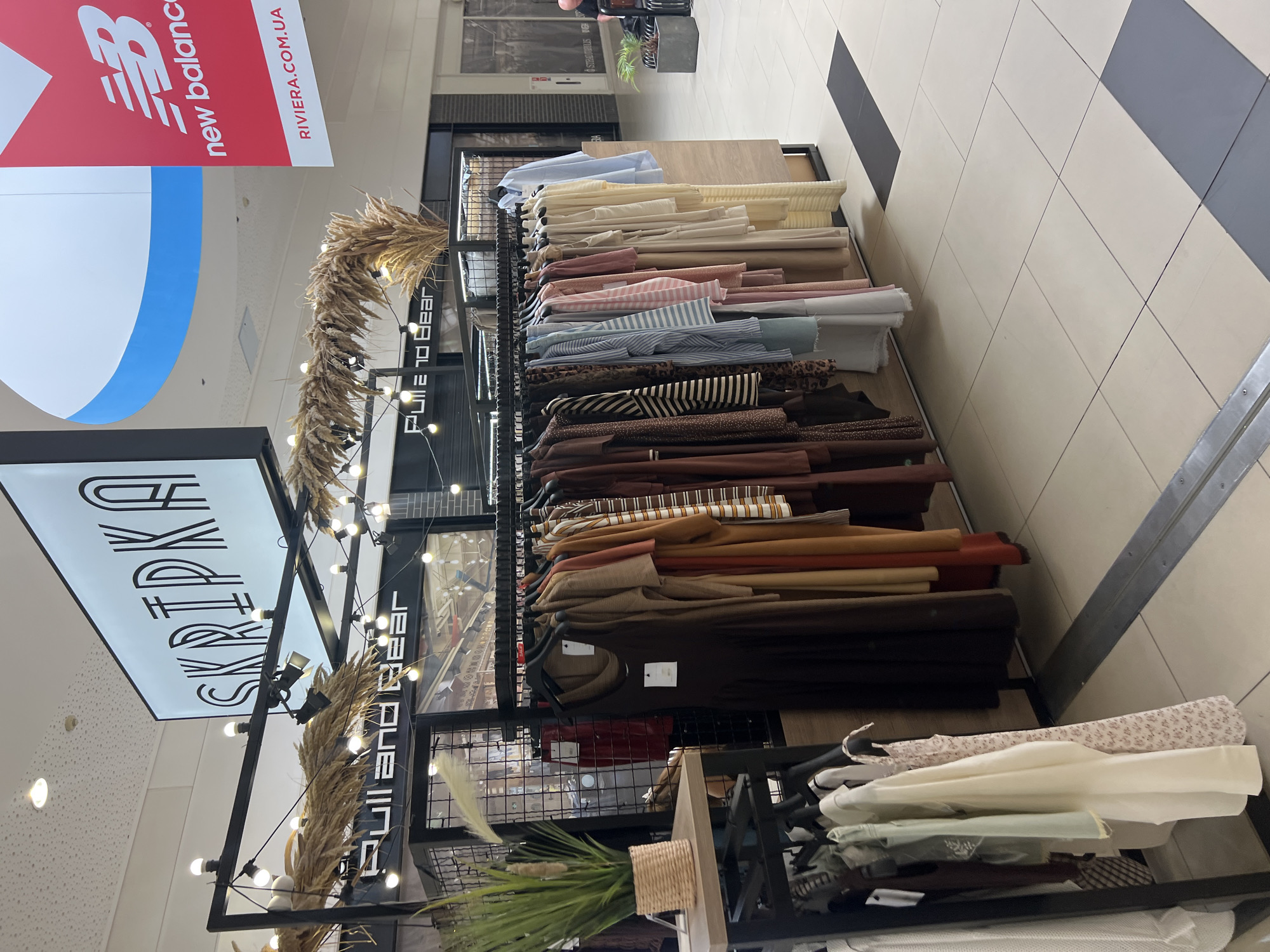 The image captures a retail display in what appears to be a shopping mall, possibly located in Fontanka, Ukraine, judging by the "RIVIERA.COM.UA" sign which is a reference to the Riviera Shopping Mall in Fontanka. The foreground features a clothing rack laden with various garments, including dresses and tops, predominantly in earthy tones and pastel colors. Some of the clothing has printed patterns, such as leopard print and floral motifs. Behind the clothing rack, a sign with stylized lettering reads "SKRIPKA". Further in the background, other retail signage is visible, including a large red "new balance" logo and a sign that reads "Pull and Bear". The environment appears to be a well-lit, modern indoor shopping space with tiled flooring. There are no people prominently featured in the immediate foreground. The overall scene depicts a commercial setting focused on fashion retail.