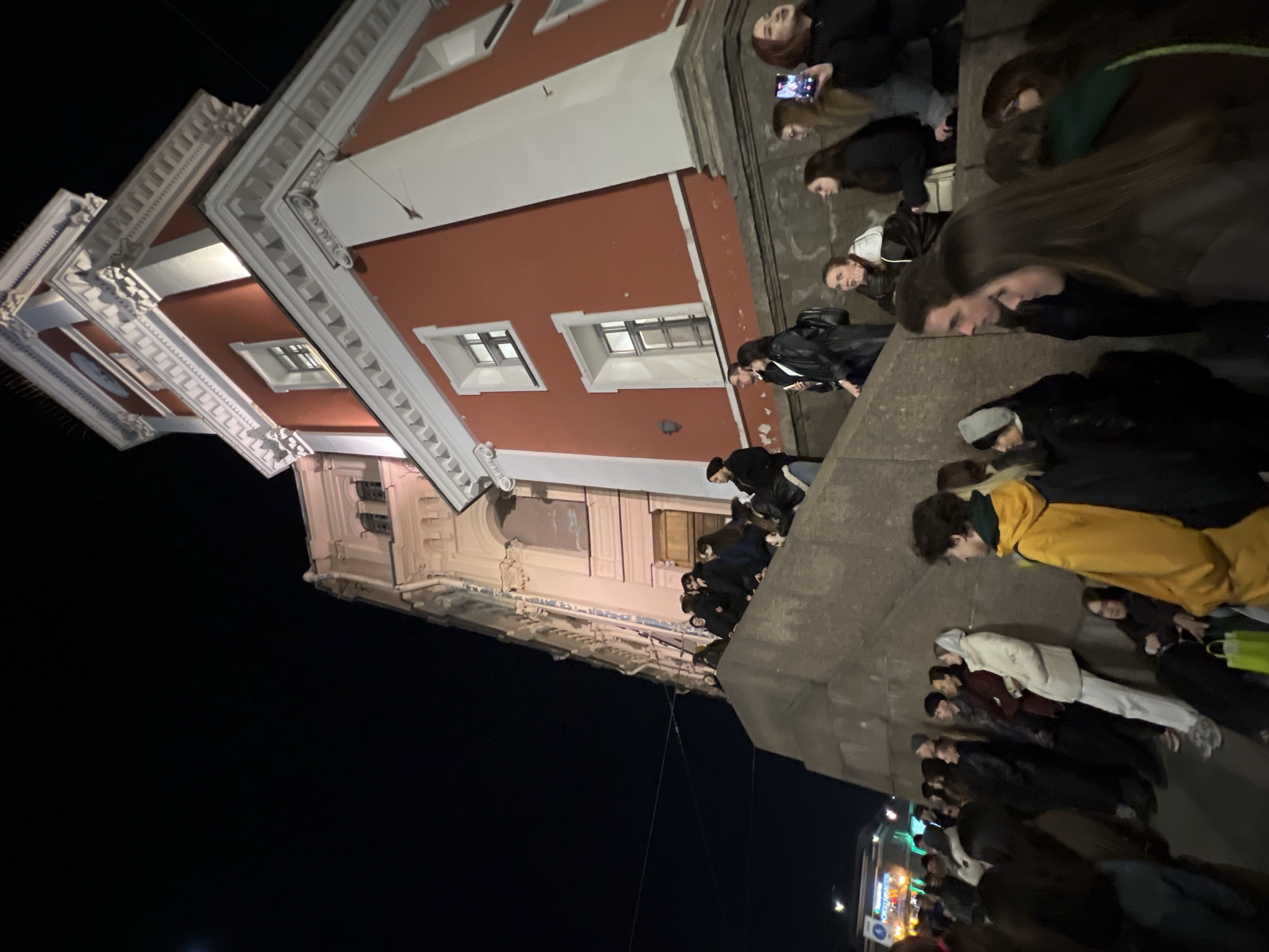 At night in Saint Petersburg, Russia, a crowd of people is gathered on the steps of a red and white building. Some individuals are standing, while others are sitting on the steps. The building is illuminated, and its architectural details, including windows and ornate cornices, are visible. The sky is dark, and a streetlight illuminates the scene at the bottom of the frame.