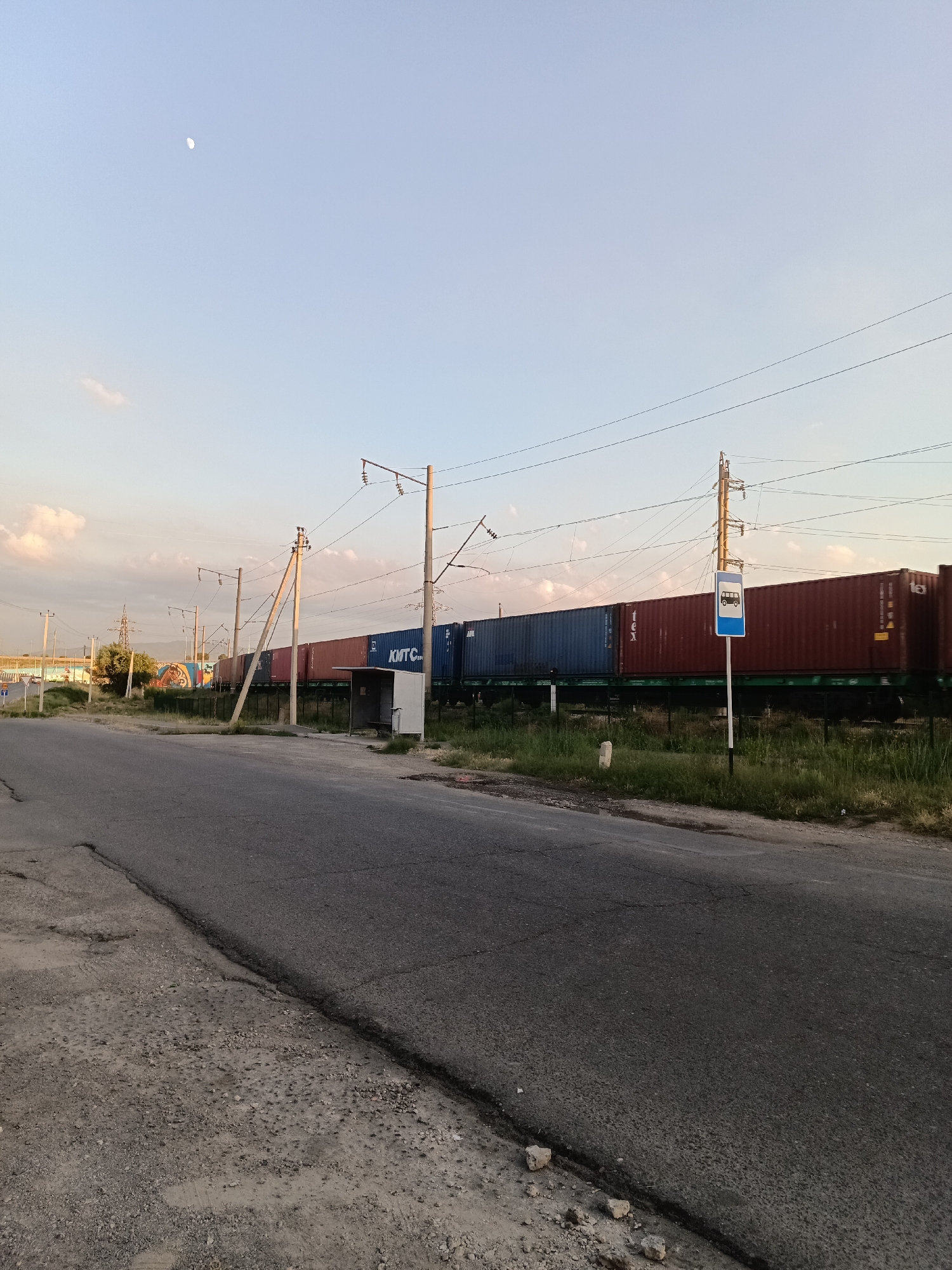 A low-angle, eye-level shot captures a freight train with several shipping containers parked along a railway track. The train is a mix of blue and red containers, some with white lettering that reads "KMTC". A small, white bus stop shelter with a blue sign displaying a white bus icon stands near the train.
The scene is set in Shymkent, Kazakhstan. A paved road runs across the foreground, with a rough, unpaved shoulder. Utility poles and overhead wires stretch across the frame, indicating an area with both road and rail infrastructure. In the distance, a cluster of trees and some low-rise buildings are visible. The sky is a pale blue with a few wispy clouds, and a sliver of the moon is visible in the upper left quadrant. The lighting suggests it is either early morning or late afternoon, with the sun casting a soft glow over the landscape. There are no people visible in the image.