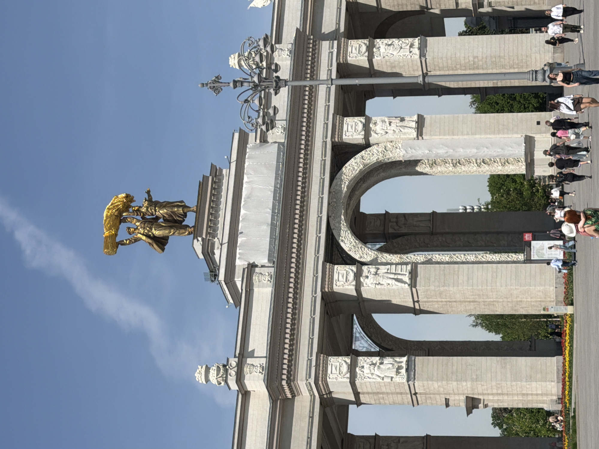 The image shows the Triumphal Arch in Moscow, Russia, from a low-angle perspective. The monument's ornate facade, topped with a golden sculpture of a chariot being pulled by horses, is prominent. The archway itself is visible, with decorative carvings. A lamppost stands near the arch. People are seen walking on the sidewalk alongside the monument. The sky is blue.