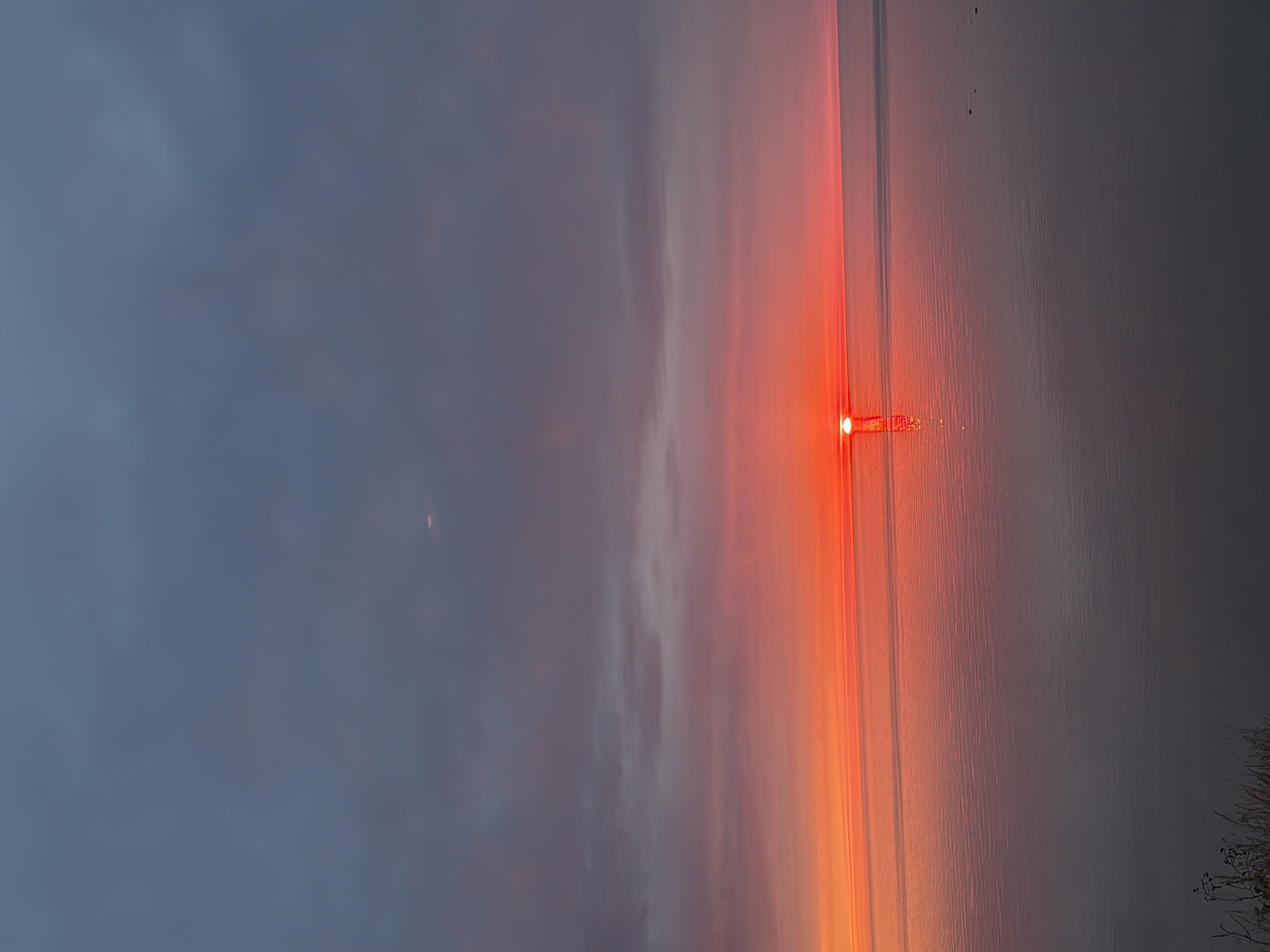 This image depicts a sunset over a calm body of water, likely the Baltic Sea, as viewed from Keila-Joa alevik, Estonia. The sun is a vibrant red orb, partially submerged on the horizon, casting a fiery glow across the sky and reflecting on the water's surface. The sky transitions from a deep, muted gray at the top to bands of orange and red as it approaches the horizon. The water is smooth, with subtle ripples that catch the light. In the foreground, the dark silhouette of dry grasses and some sparse vegetation is visible along the edge of the land. There are no people or discernable structures in the frame, creating a serene and natural landscape.
