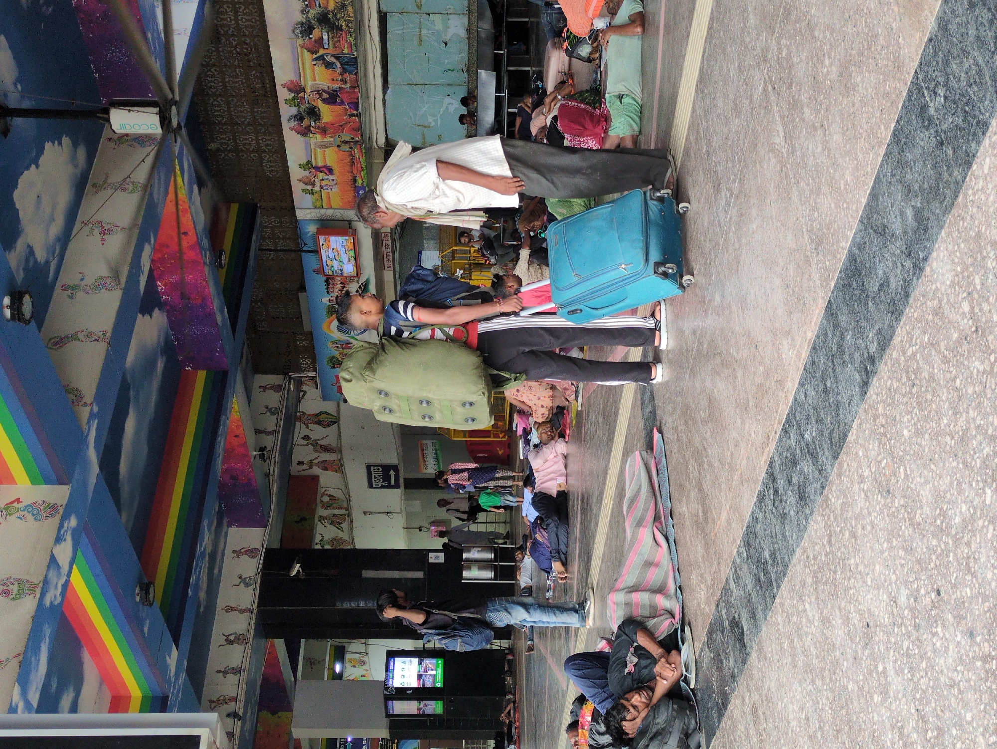 The image depicts a busy interior scene, likely a train station or bus terminal in Delhi, India, given the context provided by the prompt. The ceiling is vibrantly decorated with a rainbow motif and images of clouds and dancers. Several individuals are seen lying down on the floor, some covered with blankets, suggesting a resting or waiting area. In the foreground, a young person with a large backpack and a blue suitcase stands, interacting with an older person wearing a white shirt and scarf. Other people are scattered throughout the scene, some standing, others sitting or lying down. The floor is tiled with a speckled pattern. Information screens and murals adorn the walls, contributing to the bustling atmosphere. The overall impression is one of a public transit hub with a significant number of people either traveling or seeking shelter.