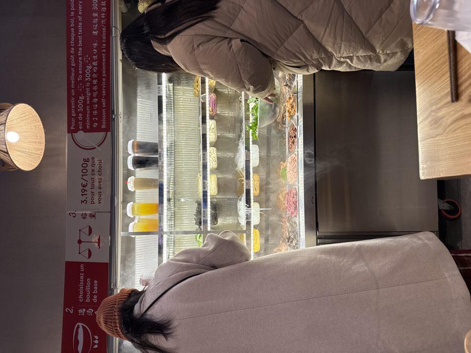 Two people are shown standing in front of a refrigerated display case with numerous compartments filled with various food items. They appear to be selecting ingredients, possibly for a self-service soup or noodle dish. A sign in French and Chinese indicates the pricing structure (€3.19 per 100g) and a minimum weight of 300g to ensure the best flavor. A lamp with a woven shade is on the left, and part of a table with a pair of chopsticks can be seen on the right.