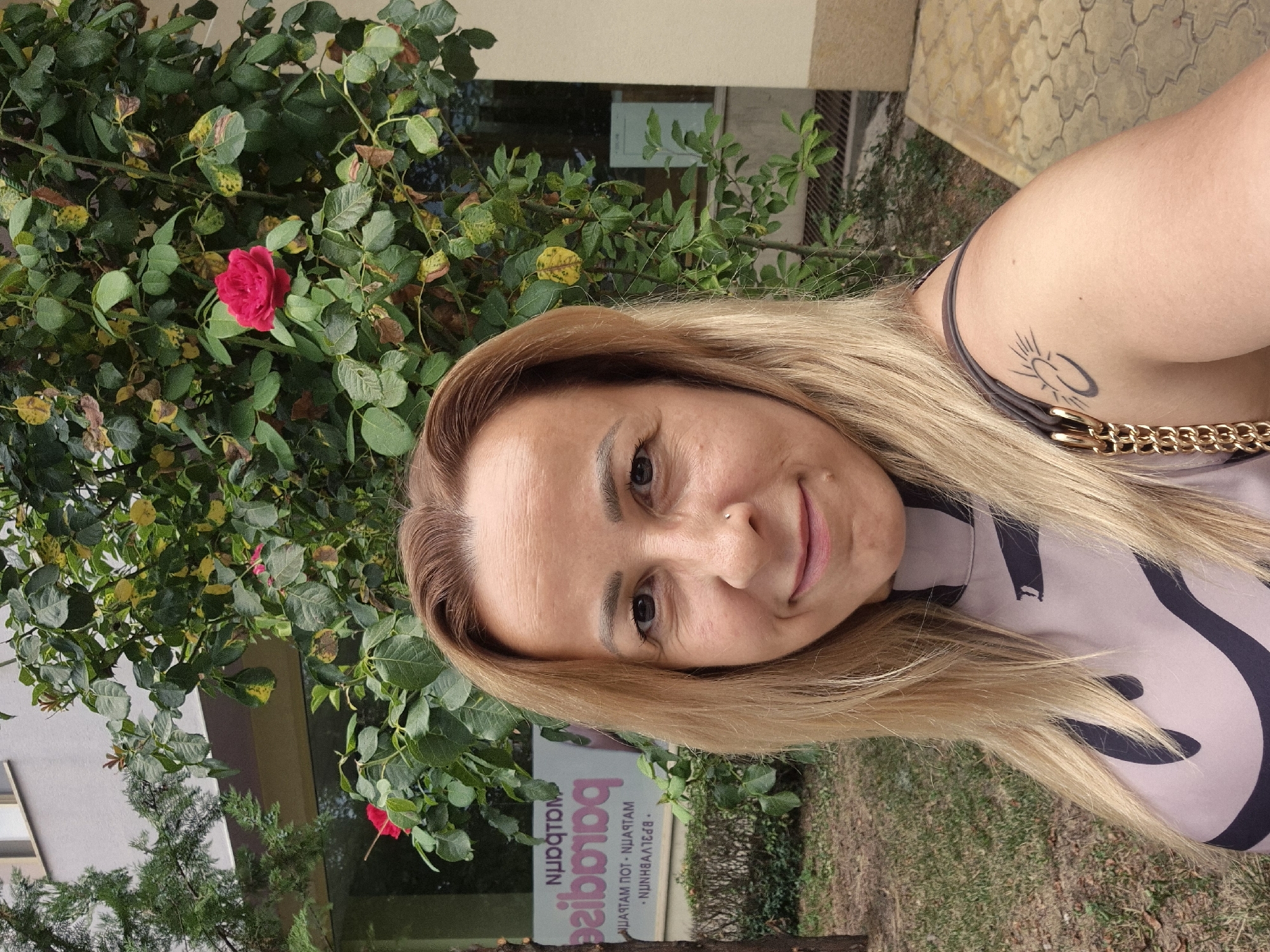 A blonde woman is taking a selfie outdoors. She is wearing a light-colored top with a dark abstract pattern and a dark purse strap across her shoulder. A tattoo of a sun with rays and a crescent moon is visible on her left shoulder. She has blonde hair, brown eyes, and a small nose ring. She is smiling slightly. Behind her is a rose bush with red roses and green leaves, a tree, and a building with windows. A sign with reversed text is visible on the building. The ground is covered with grass and dirt. The lighting suggests it is daytime.