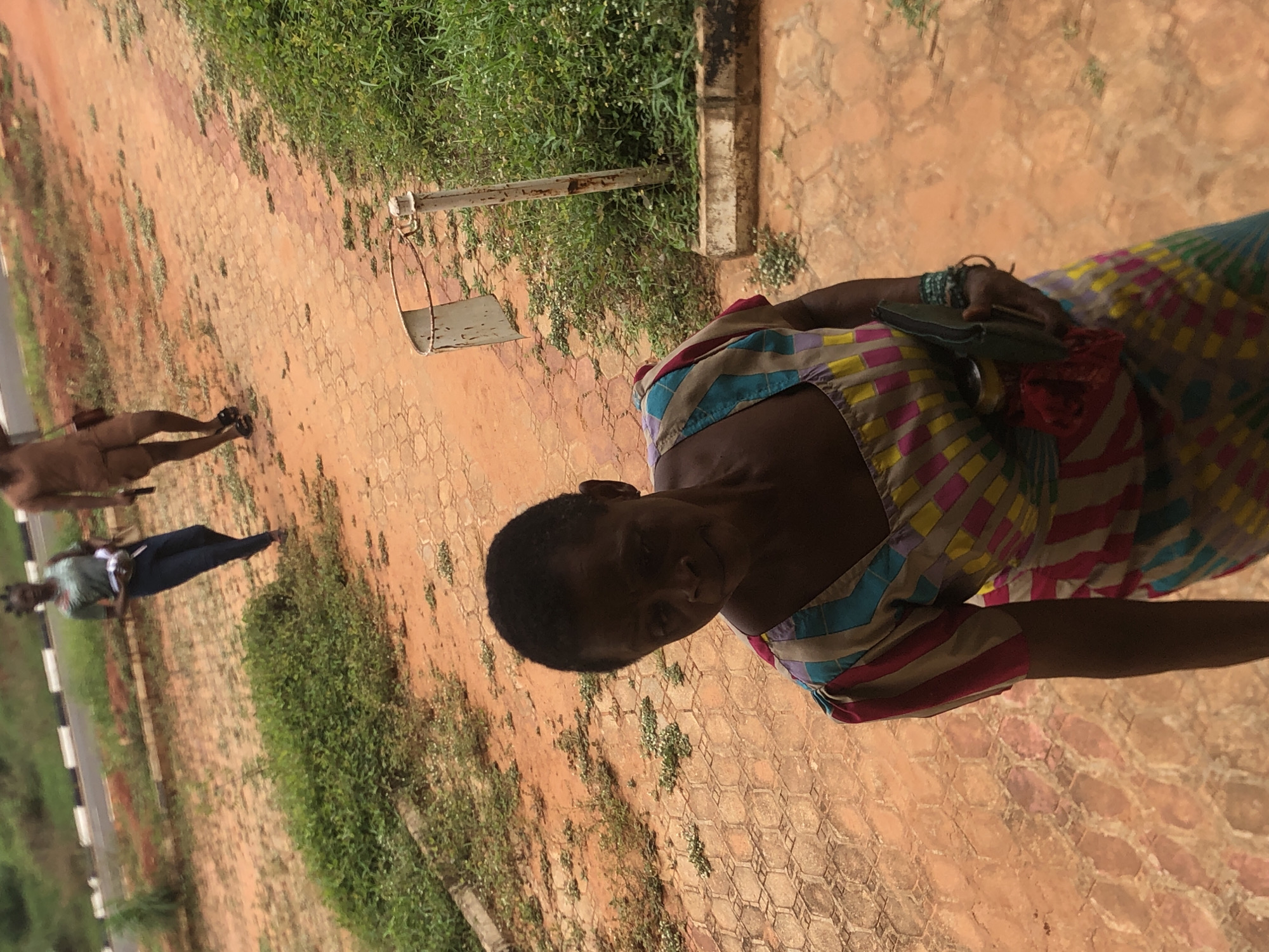 This outdoor, vertically oriented image captures a scene in what appears to be a paved area, likely in Nsugbe, Nigeria, given the provided context. The time of day is clearly daytime, suggested by the bright, natural lighting and shadows.
The primary subject is a woman positioned in the foreground, looking directly towards the camera with a neutral to slightly smiling expression. She has short, dark hair. She is wearing a sleeveless dress or top with a vibrant, multicolored striped and patterned design, featuring shades of pink, yellow, blue, and brown. On her left wrist, she wears multiple bracelets. She holds a small, dark object, possibly a phone or wallet, in her right hand, with a red fabric detail visible near it.
Behind the main subject, the ground is paved with hexagonal or interlocking reddish-brown pavers, interspersed with patches of green grass and weeds. In the mid-ground, there are two additional figures, both partially obscured and out of focus. One figure, to the left, appears to be a person standing, wearing lighter colored clothing on top and dark pants. Further back and to the right, another person is seen standing or walking.
To the right of the main woman, behind her, there is a metal structure, possibly part of a fence, gate, or playground equipment, showing some signs of rust. The background features more paved ground and greenery, with a hint of a road marked with white and black stripes in the far distance on the left side. No visible text is present in the image.