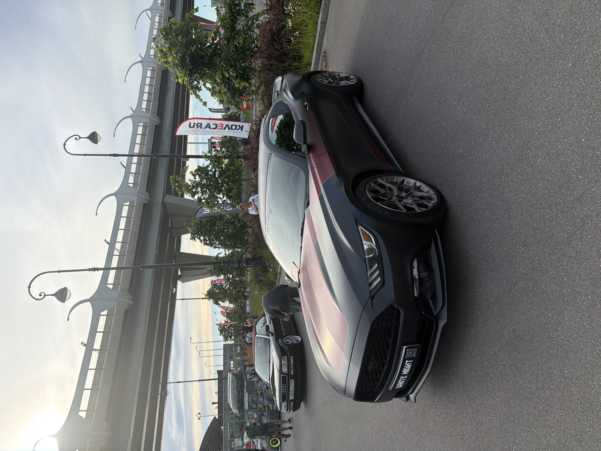 The image shows a black Ford Mustang with red racing stripes parked on asphalt, likely at an outdoor event. In the background, an elevated highway structure is visible, along with trees, flags, and other cars, including a black Audi convertible. Several people are present, some standing near the cars and others walking in the background. The lighting suggests it is daytime, possibly late afternoon. The overall scene depicts a car show or exhibition in an urban or suburban setting.