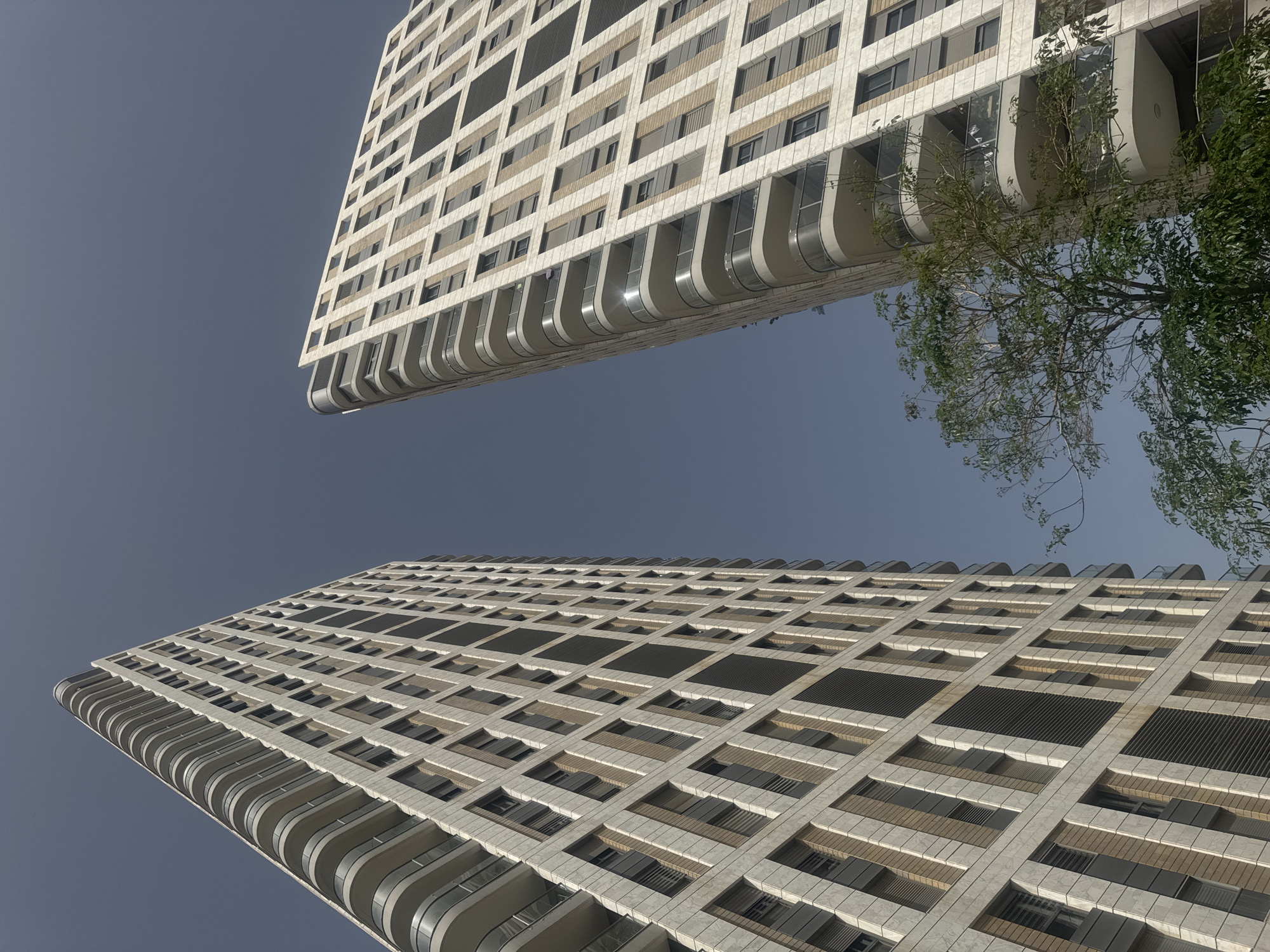 The image shows two modern, high-rise residential buildings in Givatayim, Israel. They are constructed of light-colored materials with a grid-like facade and curving balconies. The perspective is from below, looking upwards at an angle, with a light gray-blue sky visible between the buildings. A tree with sparse foliage is visible between the buildings on the right side. The architectural style is contemporary, and the buildings dominate the view.