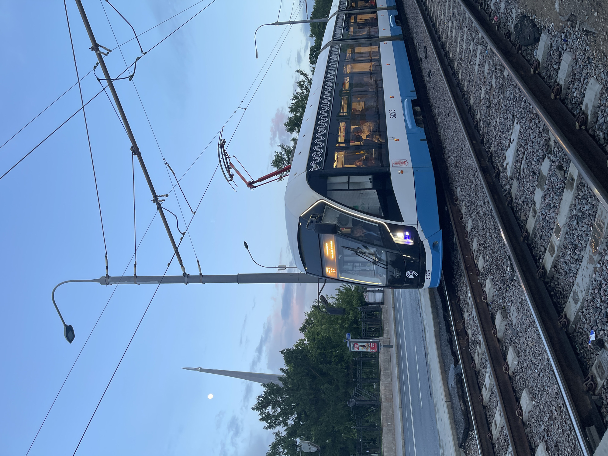 The image shows a blue and white tram on a railway track. The tram is numbered 31275. In the background, there is a tall, pointed monument. The sky is a light blue, indicating it is either early morning or dusk. There are trees along the side of the road next to the track. Overhead power lines are visible, supplying power to the tram. A street light stands near the track. The location is in the city of Unknown, Unknown.