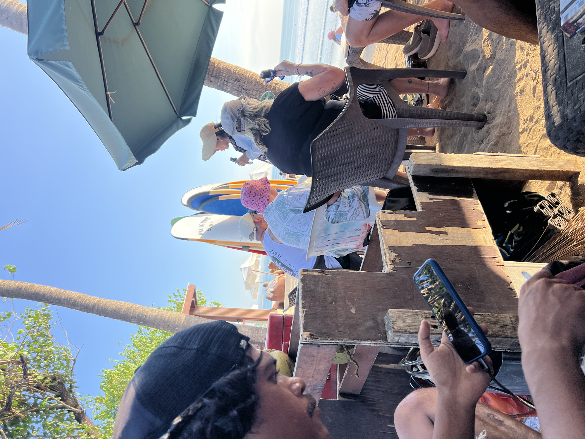 In Kuta, Indonesia, a group of people are gathered at a beachfront establishment. The scene is set on a sandy beach, with palm trees and the ocean visible in the background. An umbrella shades some of the patrons. Several surfboards are propped up nearby. In the foreground, a person wearing a black beanie is focused on their phone, which is held in their hand. Other individuals are seated at tables, some engaging in conversation and others holding drinks or food items. The overall atmosphere appears relaxed and casual, typical of a beachside resort.