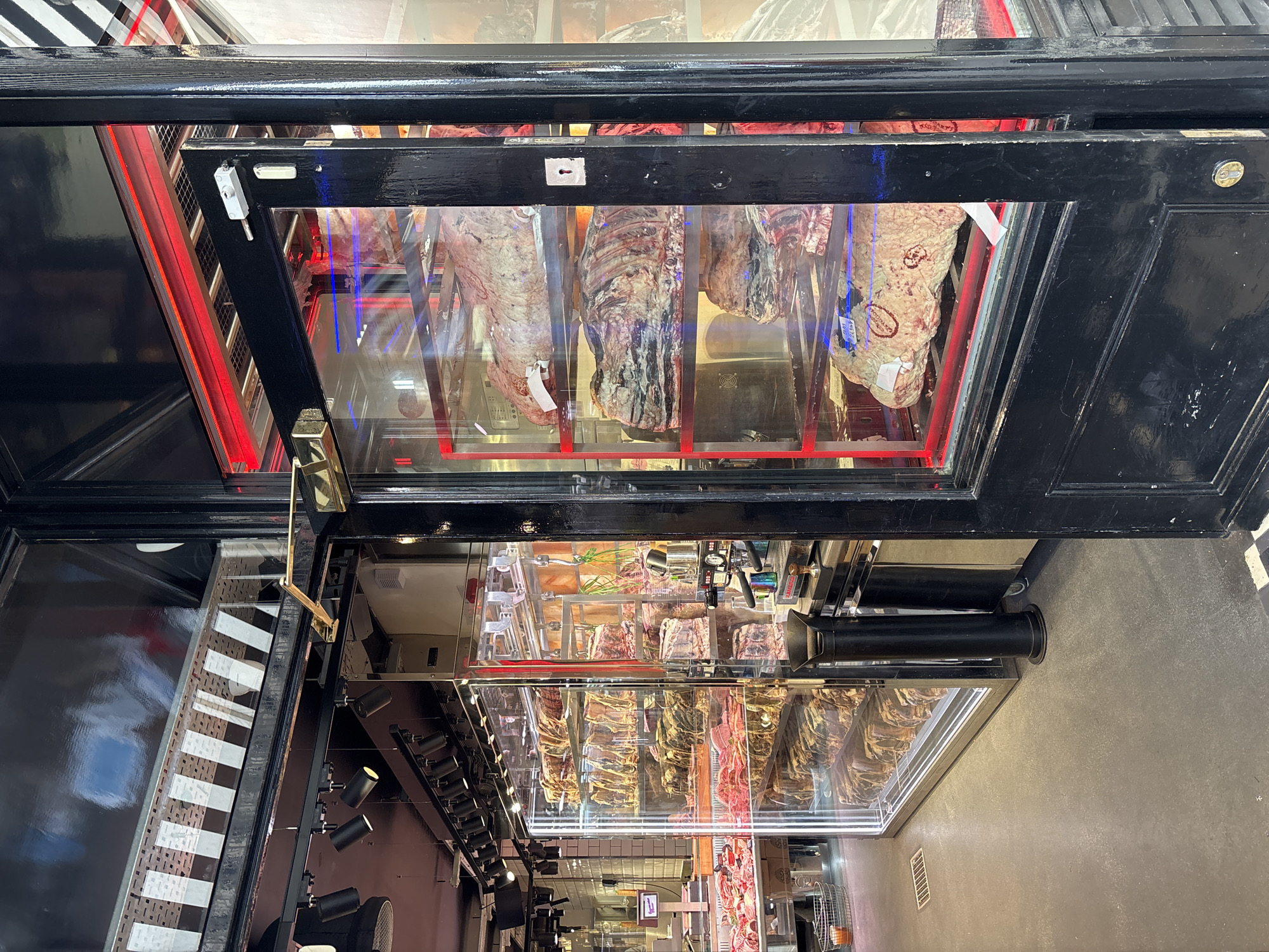The image shows a butcher shop in what appears to be a high-end area, potentially in London, United Kingdom. Several large cuts of meat hang within a refrigerated display case with a black frame. Below, a larger horizontal display case contains various cuts of meat arranged neatly. A lighting system is visible along the ceiling near the display cases. The floor is a simple concrete grey. The overall impression is of a modern and sophisticated butcher shop.