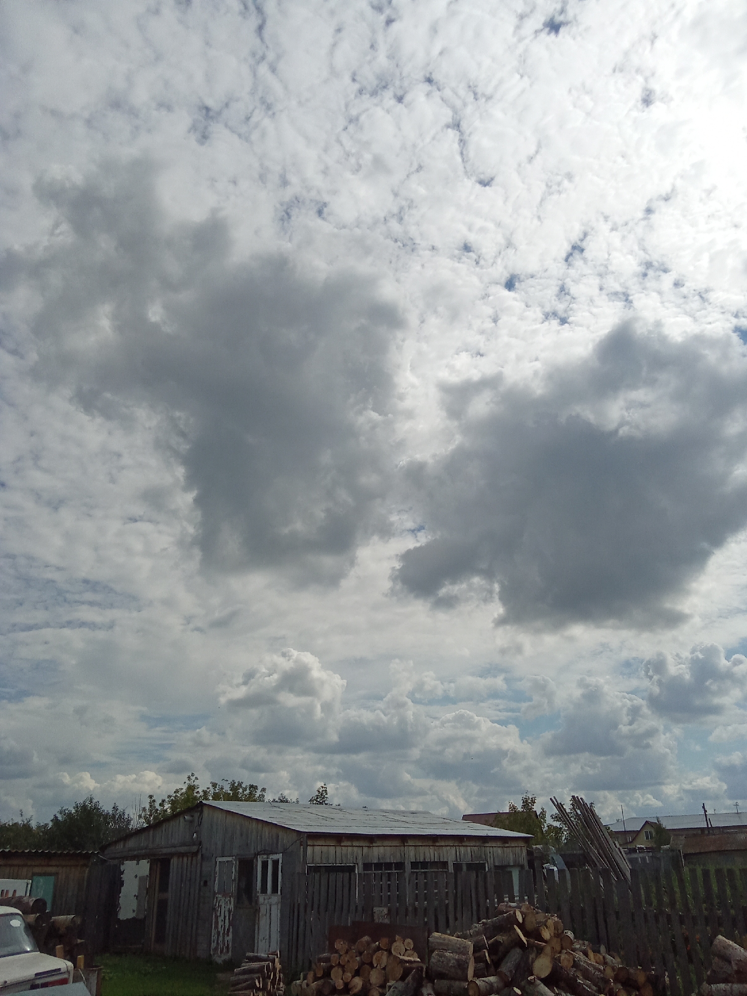 The image depicts an outdoor scene in Рощин ауылдық округі, Kazakhstan, under a cloudy sky. In the foreground, a weathered wooden shed with a corrugated metal roof is visible, featuring several doors and windows, some of which appear damaged or open. Piles of cut logs are stacked near the shed and a wooden fence. A portion of a white vehicle is also present on the left side of the frame. In the background, more wooden structures and trees are discernible, along with a glimpse of houses and a utility pole. The sky is filled with a mix of light and dark clouds.