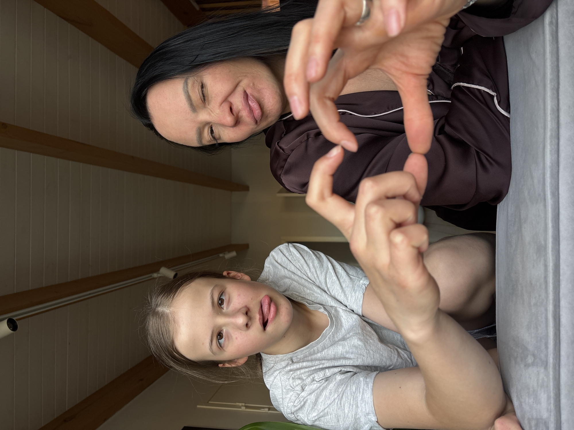 Two individuals are present in the photograph. One, appearing younger, sticks out their tongue. The other, seemingly older, is positioned above and behind the first, with their hands forming a heart shape. The background reveals an interior space with wooden beams and paneling. The location is in the city of Chaikovsky, Russia.