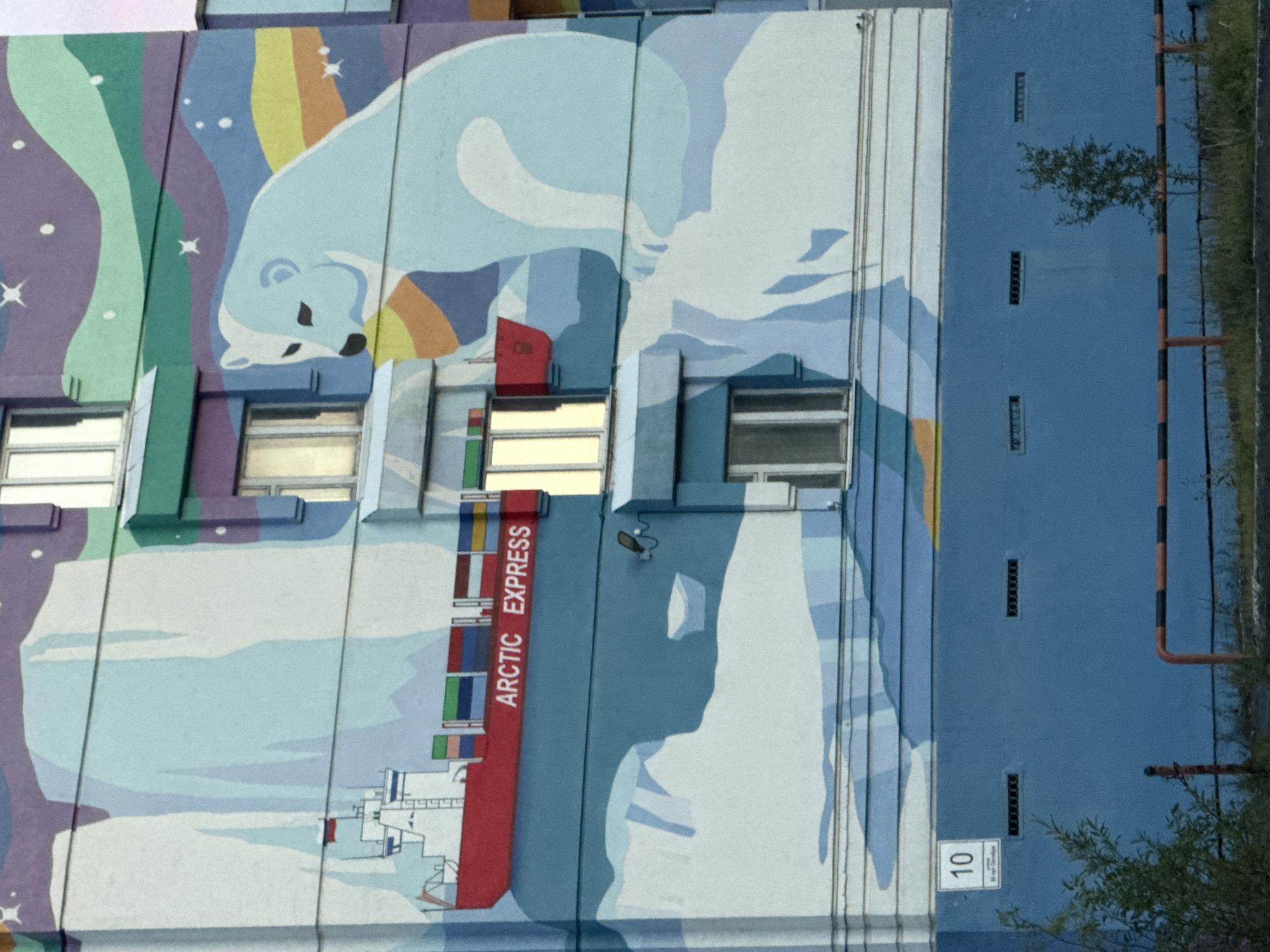The image is a photograph of a mural painted on the side of a multi-story building in Norilsk, Russia. The mural depicts an Arctic landscape with a polar bear, icebergs, and a cargo ship labeled "ARCTIC EXPRESS." The building itself is painted in a pale blue color at the lower section, with windows visible on multiple floors. The upper sections of the building are covered by the mural, which features a stylized aurora borealis in shades of purple, green, and yellow. The polar bear is light blue and appears to be sitting on an iceberg. The cargo ship is red and is depicted in the water among ice floes. The building has a sign with the number "10" on its lower blue section. In front of the building, there is a metal barrier with orange and black stripes, grass, and a small tree. The overall scene is a colorful and artistic representation of an Arctic environment on an urban structure.