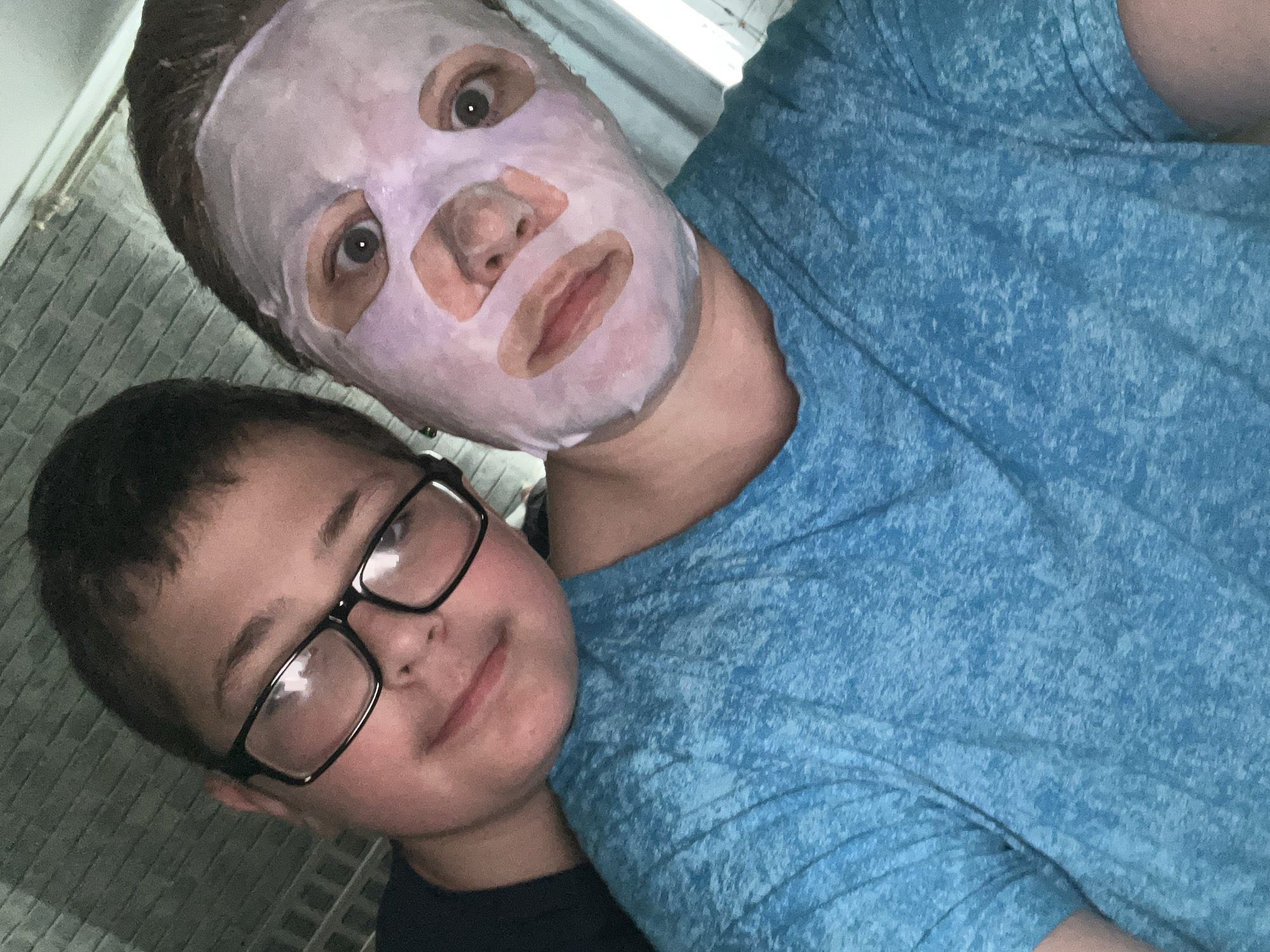 A low-angle, selfie-style photograph shows two individuals. One, a person wearing a lavender-tinted facial mask with cutouts for the eyes, nose, and mouth, is positioned in the upper portion of the frame. Their eyes are wide, and their expression is neutral. The second person, a child with dark hair and glasses, is leaning against the first person, occupying the lower left portion of the image. The child is looking at the camera and smiling slightly. The first person is wearing a blue t-shirt. A partially visible brick wall is in the background. The location appears to be indoors. There is no visible indication of Vesyoloye, Russia.