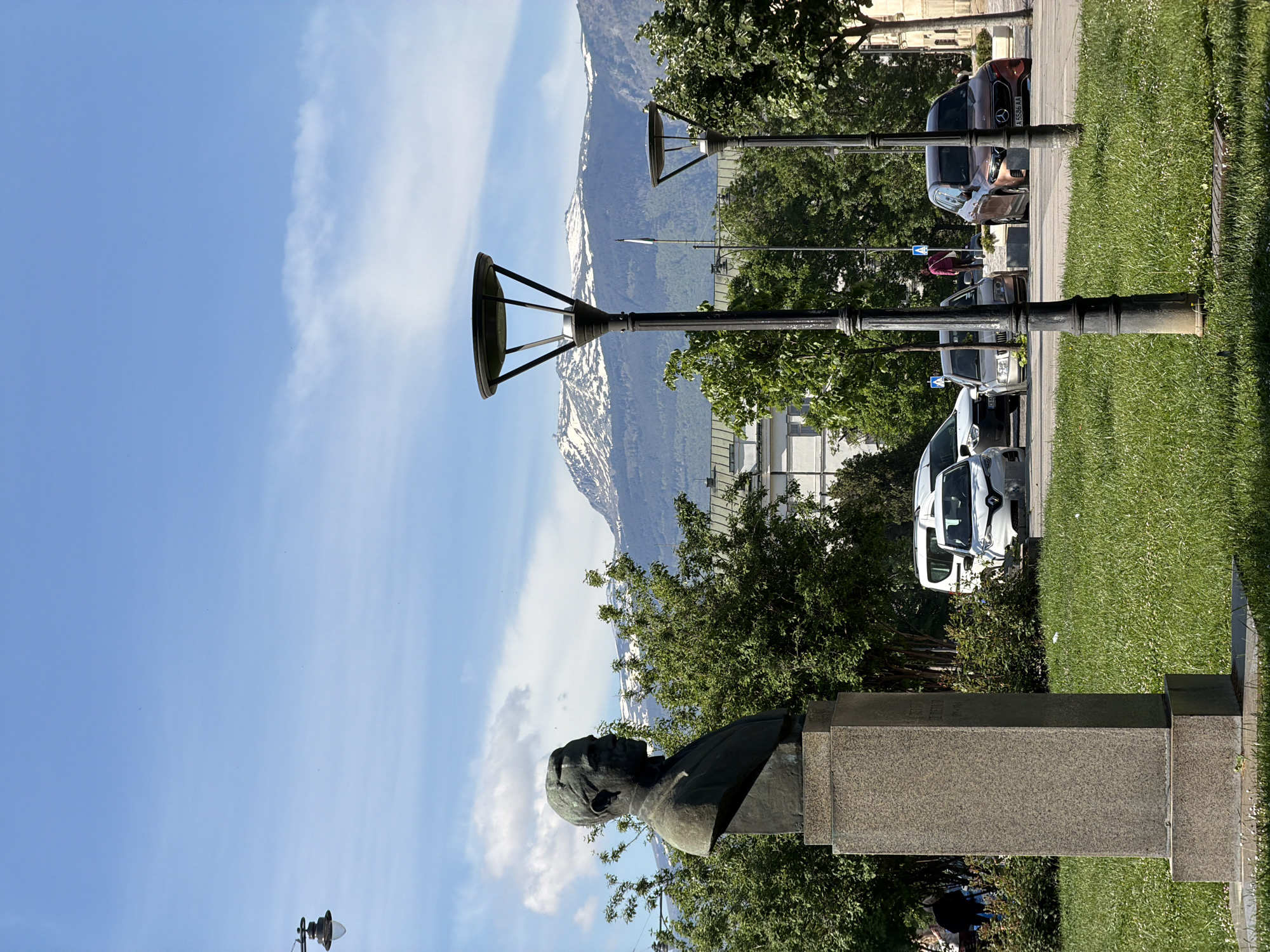 The image depicts a street scene in Sofia, Bulgaria. A bronze bust atop a stone pedestal stands in a green park area, with a mountain range visible in the background. Several street lamps line the sidewalk, and parked cars are seen along the curb. There are people near the cars. The sky is blue with some white clouds.