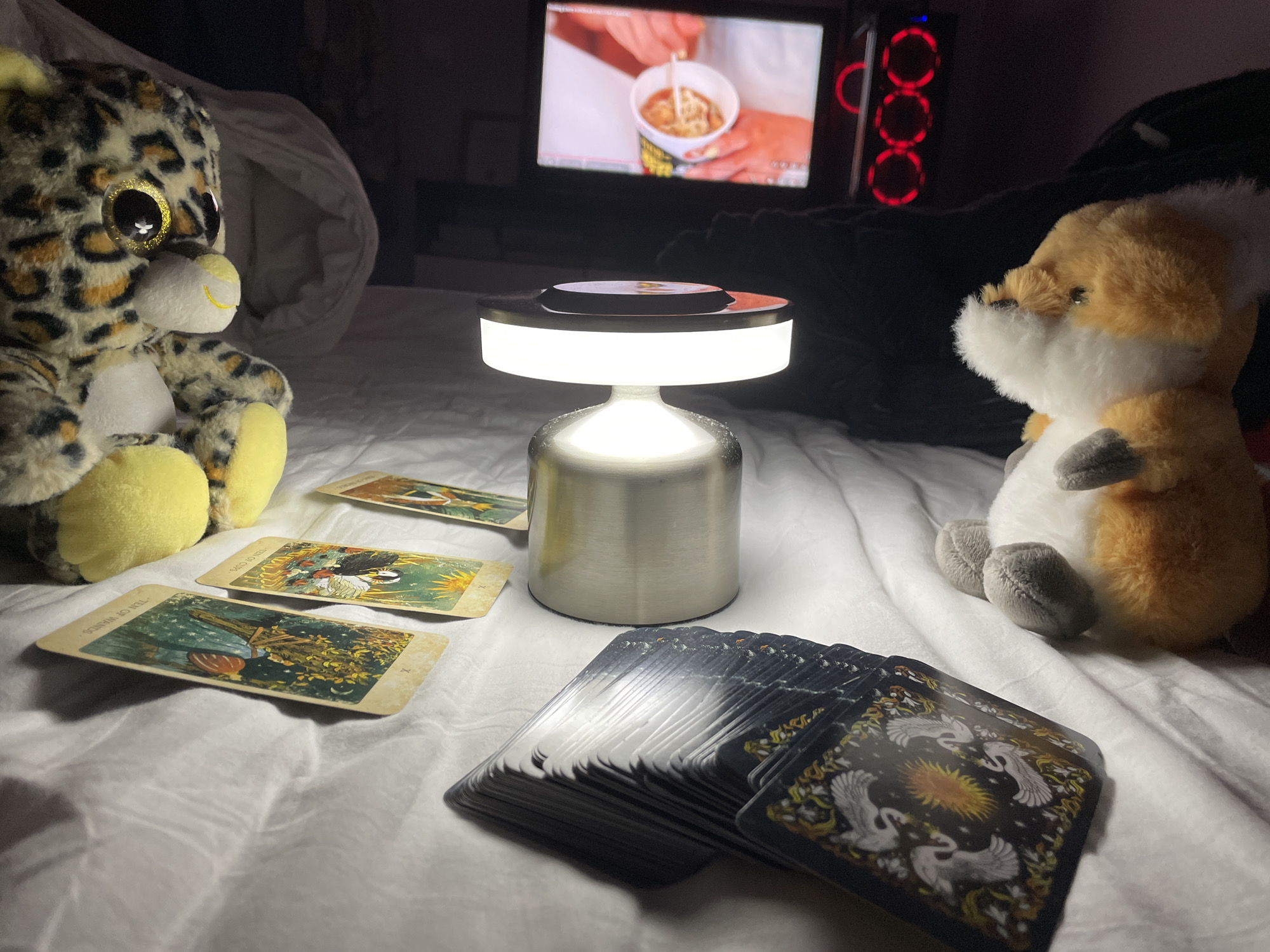 The image shows a dimly lit bedroom scene, possibly in Veliko Tarnovo, Bulgaria, though there is no direct evidence of this location in the photograph. On a white bedspread are a deck of tarot cards and three cards laid out in a spread. A small silver lamp illuminates the scene. A stuffed leopard and a stuffed fox are positioned on either side of the arrangement, seemingly observing the cards. In the background, a television screen displays an image of someone holding a cup of noodles. Next to the television is a computer tower with red LED rings.