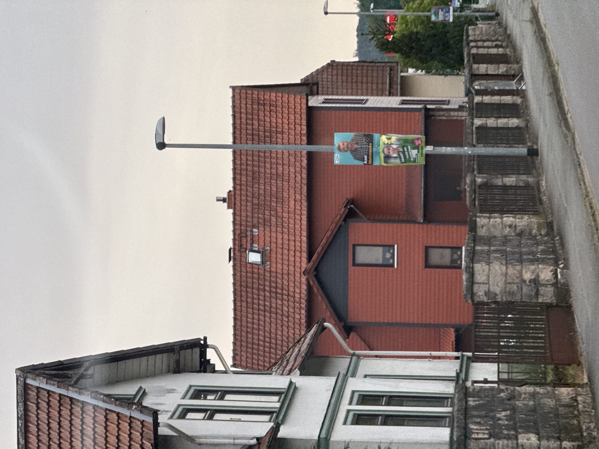 The image shows a street scene in Bad Sachsa, Germany. A red residential building with a tiled roof is prominently featured. A lamppost stands in front of the building, displaying two political posters. The top poster features a man in a plaid shirt, identified as Axel Mehnecke, with the "CDU" logo visible. The lower poster shows a young person with a sunflower. A white house with dark trim and a tiled roof is partially visible to the left. In the foreground, a stone wall with a metal fence runs along the sidewalk. The sky is overcast. In the background, trees and distant lights are visible.