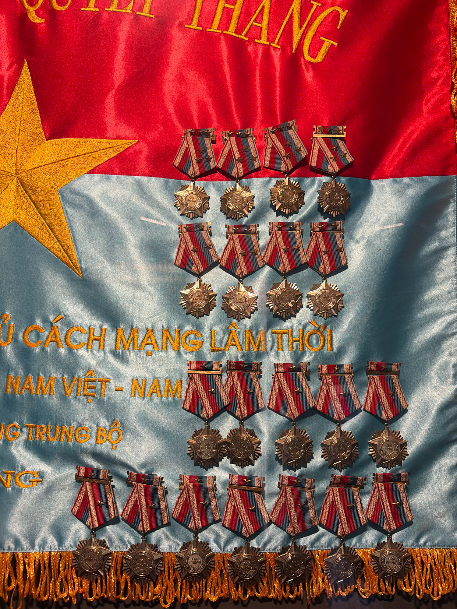The image shows a close-up of a red and light blue flag with gold embroidery and several medals hanging from it. The flag features a large gold star on the left side and text embroidered in gold. The text appears to be in Vietnamese. The medals are of various sizes and designs, with ribbons in red, white, and blue. The lower edge of the flag has gold fringe. The medals and flag suggest a patriotic or military context. The provided information about the location in Đà Nẵng, Vietnam, is external to the visual content of the image.