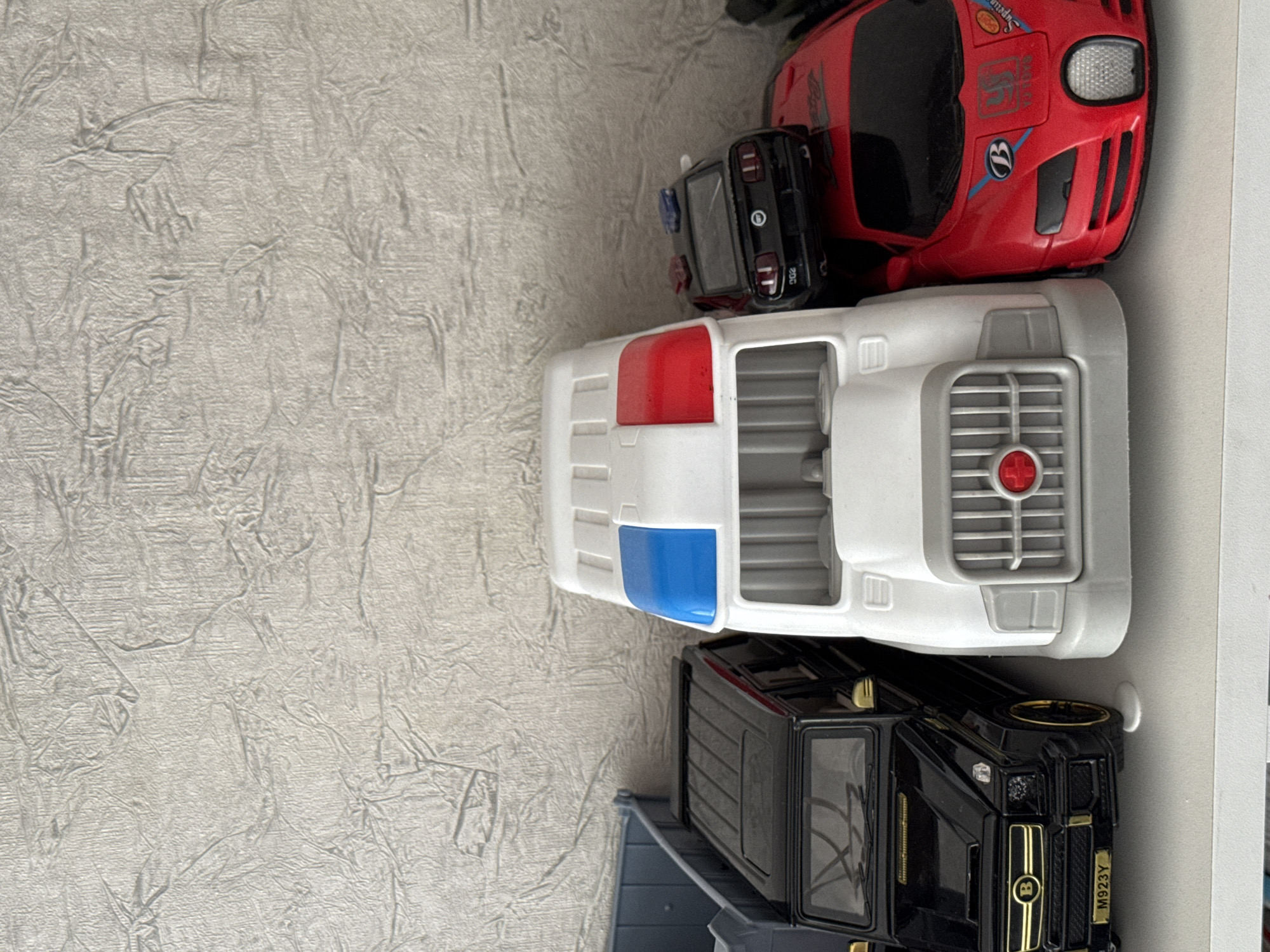 A collection of toy vehicles is displayed on a white shelf against a textured, light-colored wall. From left to right, the first visible toy is a black SUV, resembling a Mercedes-Benz G-Class, with gold accents and a license plate that reads "M923Y". To its right is a large, white toy truck with blue and red lights on its roof. Stacked on top of the white truck is a black toy police car, a Ford Mustang, with blue and red emergency lights. Next to the white truck and partially obscured by it is a red toy sports car, resembling a Bugatti, with various decals and markings. The background wall has a repeating pattern of raised, irregular lines. The lighting appears to be from above, casting subtle shadows on the shelf and wall. There are no people or discernible signs of human activity in the immediate scene. The location is specified as Yarominski Sel'ski Sovet, Belarus.