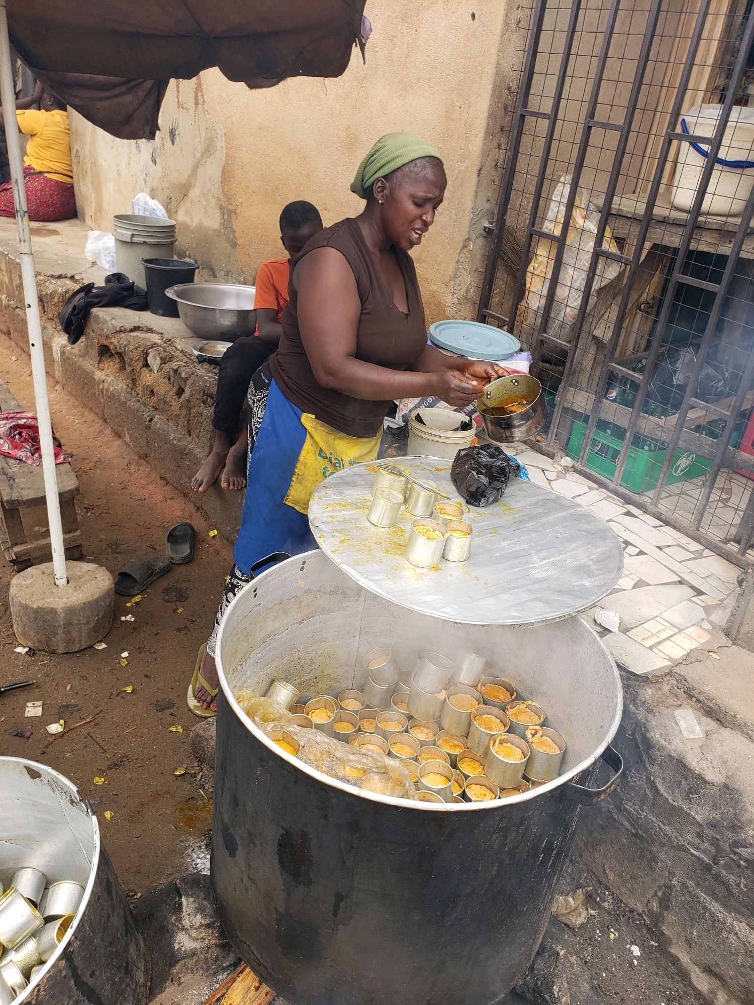 A Nigerian woman wearing a green headscarf and a brown top is standing over a large metal pot filled with food. She is holding a small pan of what appears to be oil over the pot. The pot is also filled with small metal containers, each containing food. The scene appears to be outdoors, with a rough concrete ground and a wall in the background. There are other people and objects in the background, suggesting a busy marketplace or street food stall. The overall impression is one of food preparation in a local setting in Narayi, Nigeria.