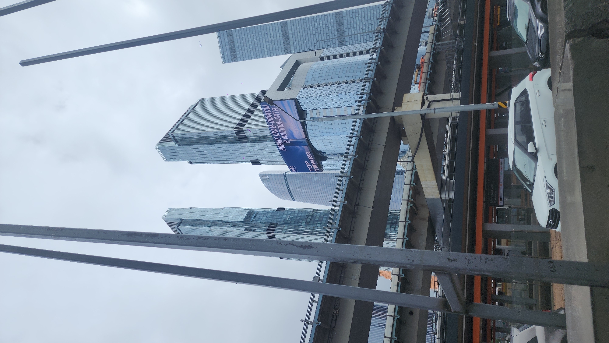 The image shows a view of the Moscow International Business Center (Moscow-City) from an elevated perspective, possibly from a bridge or overpass. Several modern glass skyscrapers dominate the skyline. A large advertising banner is attached to one of the buildings. A road or transit line runs through the scene, with cars visible on the right side of the frame. The sky is overcast. Structural elements, such as metal railings and supports, are in the foreground.