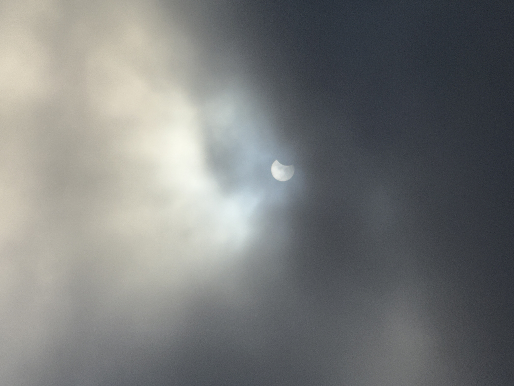 A partial solar eclipse is visible through dense, overcast clouds in Vila Nova de Gaia, Portugal. The sun, partially obscured, appears as a crescent shape with a distinct bright edge peeking through a gap in the dark, textured clouds. The sky is dominated by varying shades of gray and white, with a luminous glow emanating from behind the clouds where the sun is positioned. The overall lighting is dim and diffused, typical of cloudy weather. The image captures a specific moment of a celestial event occurring in an atmospheric, muted environment.