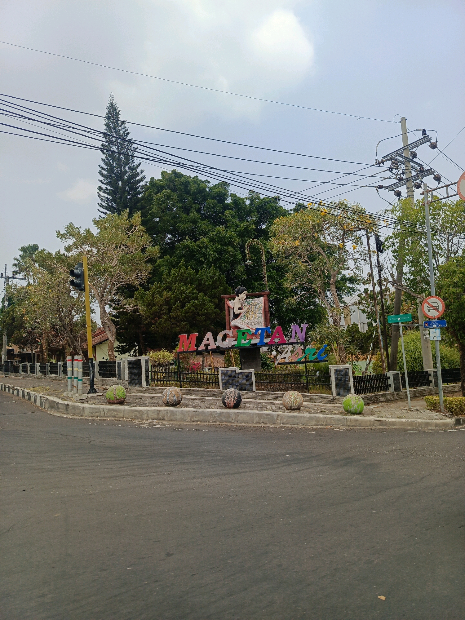 The image captures a street view in Sukowinangun, Indonesia, featuring a prominent sign that reads "MAGETAN ASRI". The sign is brightly colored and stands in front of a dense foliage of trees, with a tall pine tree visible in the background. In the foreground, a road curves around a traffic island, which is adorned with decorative spherical objects. A traffic light, a utility pole with numerous wires, and various street signs are also present. The sky is overcast, suggesting a cloudy day. No people are visible in the frame.