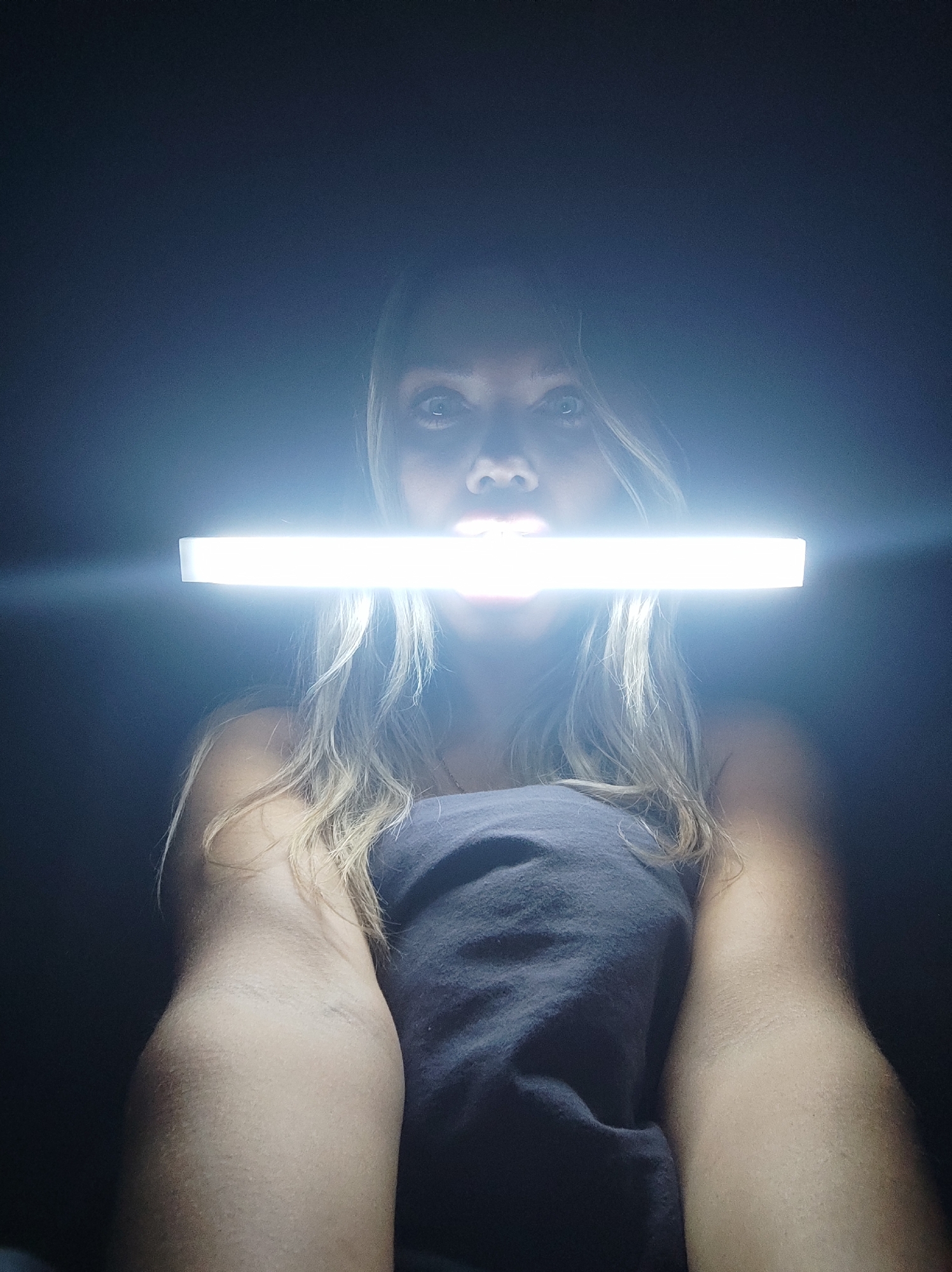 A fair-skinned woman with shoulder-length blonde hair holds a bright white rectangular light source in front of her mouth. She wears a dark, strapless top and appears to be indoors against a dark background. There is no indication of the location being Votkinsk, Russia or any other city.