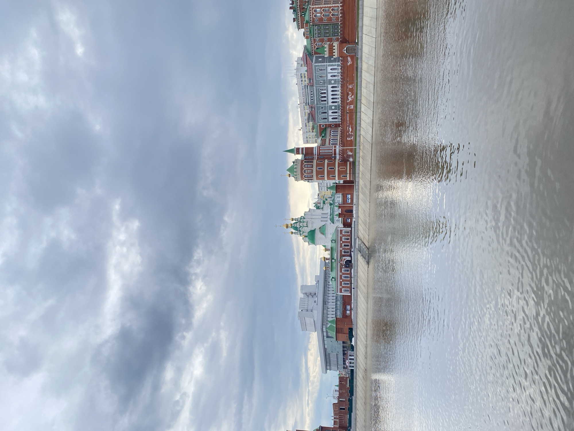 The image captures a wide-angle view of a distinctive cityscape along a tranquil waterway in Yoshkar-Ola, Russia. The scene is dominated by a river or canal in the foreground, its surface covered with gentle ripples and reflecting the soft, diffused light from the sky. A light-colored concrete embankment runs parallel to the water, forming a clear boundary between the natural element and the urban structures.
Across the embankment, a row of buildings stretches along the waterfront, showcasing a unique architectural style. Many structures are built with reddish-brown brick, contrasting with white decorative elements, window frames, and cornices. Roofs are predominantly green or gray. Several prominent buildings feature traditional Russian Orthodox architectural elements, including green-domed spires and gilded onion domes, suggesting the presence of churches or cathedrals within the complex. The architecture overall appears ornate and well-maintained, blending traditional Russian designs with European influences.
The sky above is heavily overcast, filled with thick layers of gray and white clouds, indicating a cloudy day. Despite the cloud cover, the scene is well-lit, suggesting it is daytime, likely late morning or early afternoon. No individuals or specific activities are visible within the frame, contributing to a calm and architectural ambiance. There is no discernible text in the image.